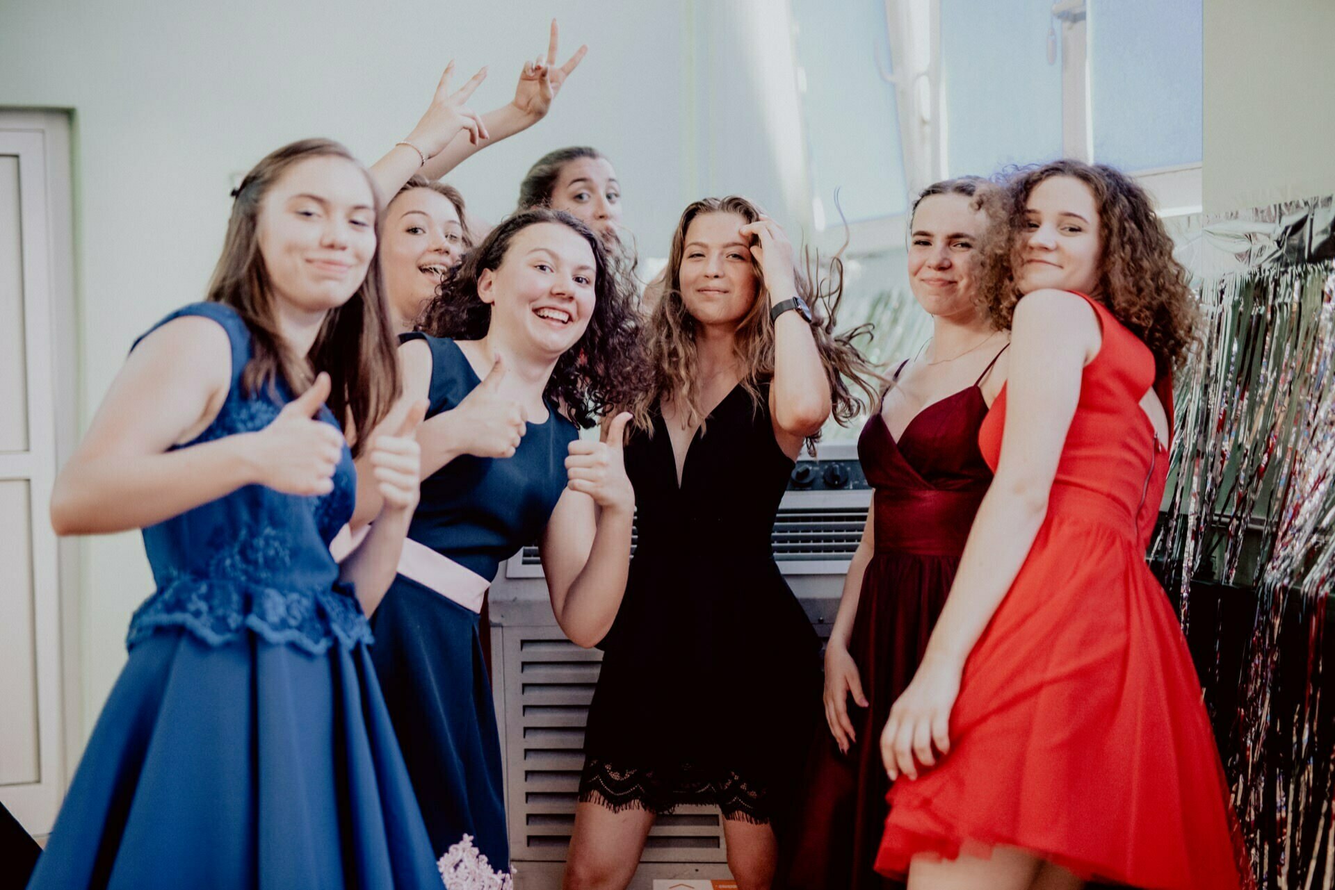 A group of seven young women in formal attire pose for the photo. They are all smiling and making various gestures such as thumbs up and peace signs. The background shows a room with bright walls and a decoration of metallic tassels, perfectly captured by the event photographer Warsaw.  