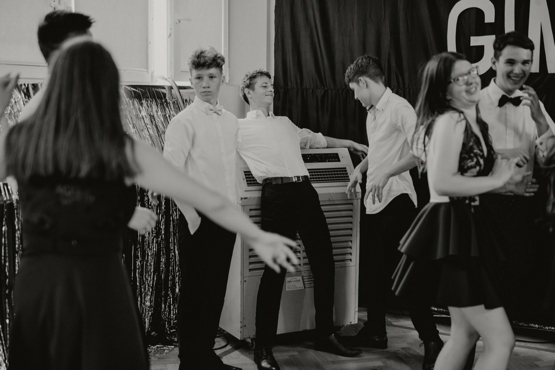 A group of young people dressed in formal attire are dancing and enjoying themselves at a party being held indoors. One of the boys is leaning dramatically against an air conditioner, surrounded by friends who are also enjoying the moment. The mood is lively and festive, which this event photography captures perfectly.  