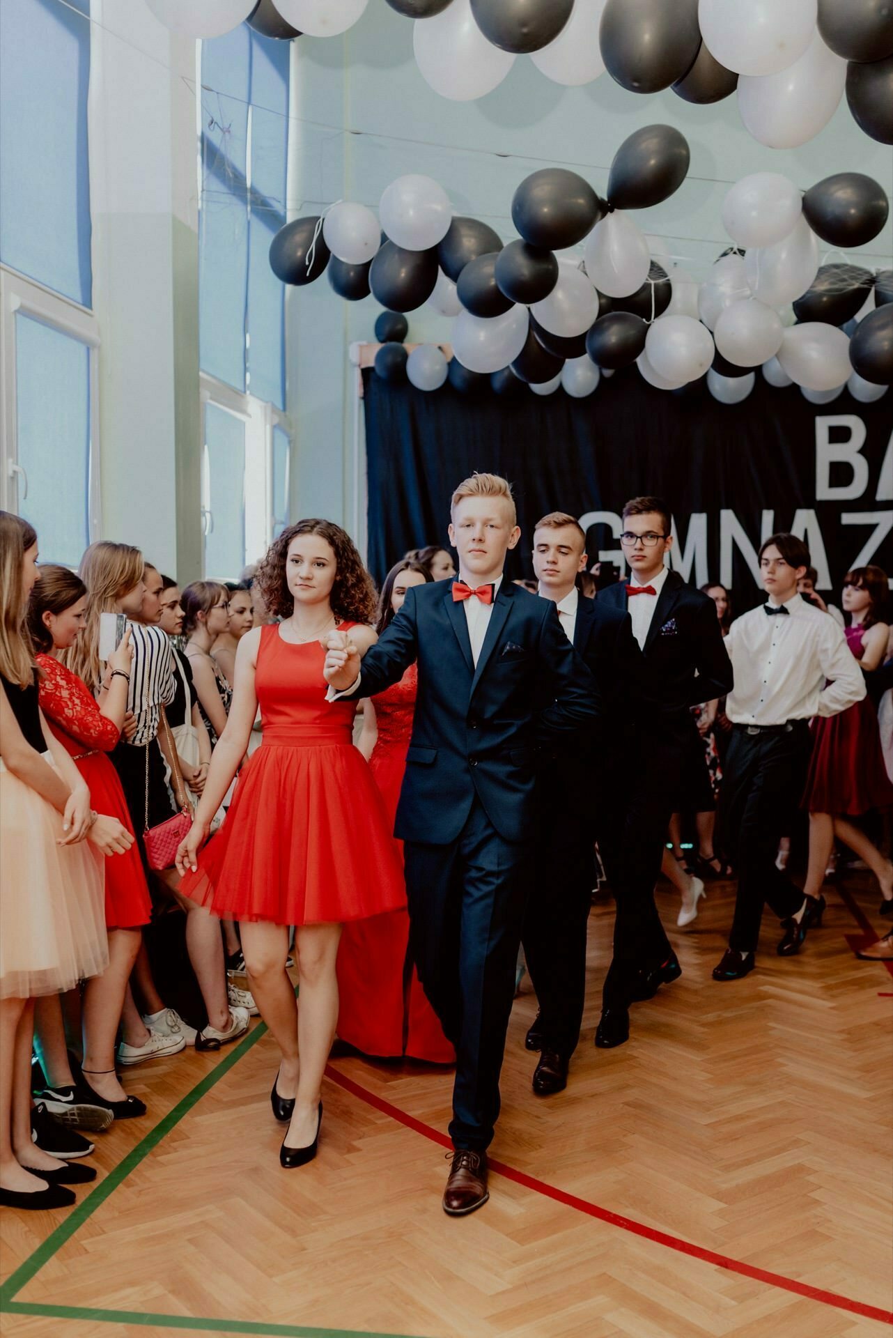 A group of young people in formal attire walk in pairs at a school dance or formal party, beautifully captured by an event photographer Warsaw. The boys are mostly dressed in dark suits with bow ties, while the girls wear a variety of dresses, including a red one that the girl wears in front. The space is decorated with black and white balloons.  