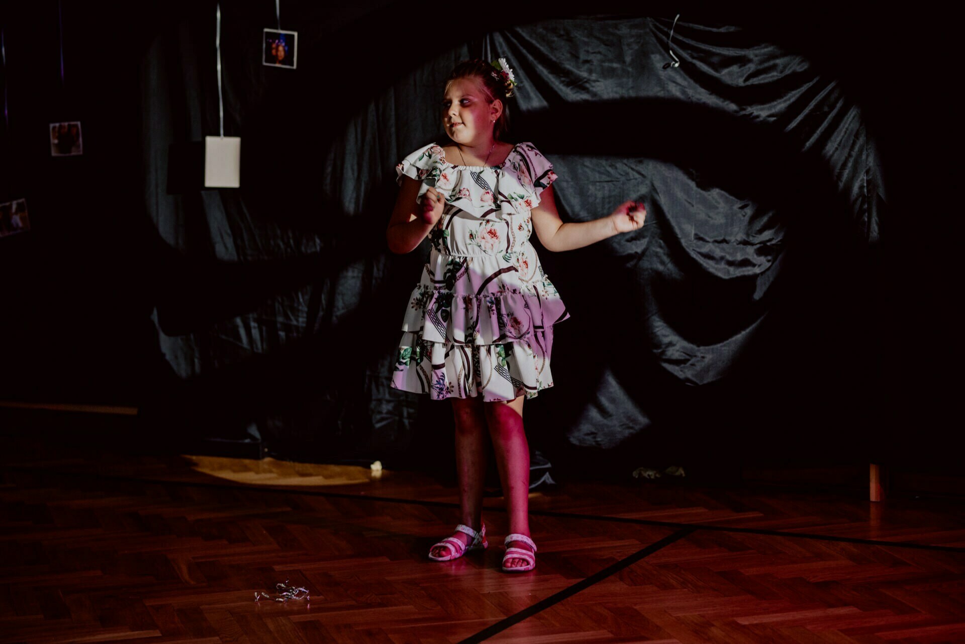 A young girl dressed in a floral dress and sandals stands on a wooden floor. She is illuminated by pinkish stage lighting, with shadows behind her on a dark background. She appears to be performing or posing in the spotlight, captured as part of an event photograph.  