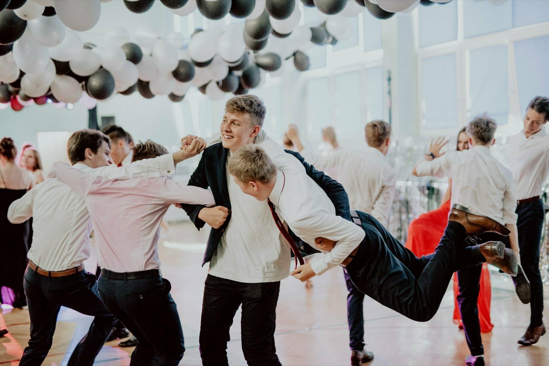 A group of young men dressed in formal attire joyfully dance and celebrate under a ceiling decorated with black and white balloons. Two other men playfully lift them from the ground. The atmosphere is lively and festive, perfectly captured in the photo report of the events.  