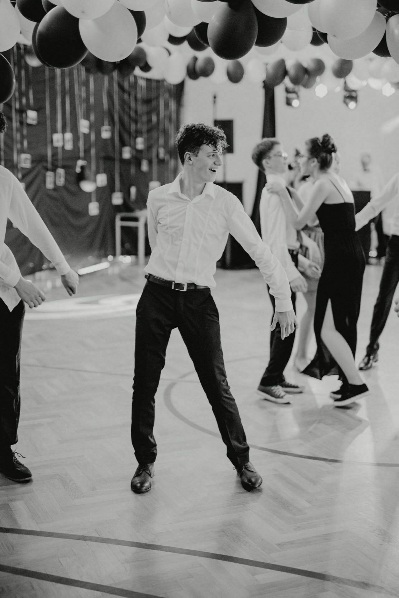 The black and white event photo shows a young man in a white shirt and black pants dancing merrily at a party. Other people are dancing in the background, with balloons hanging from the ceiling. The mood is lively and festive, perfectly capturing the event photo by event photographer warsaw.  