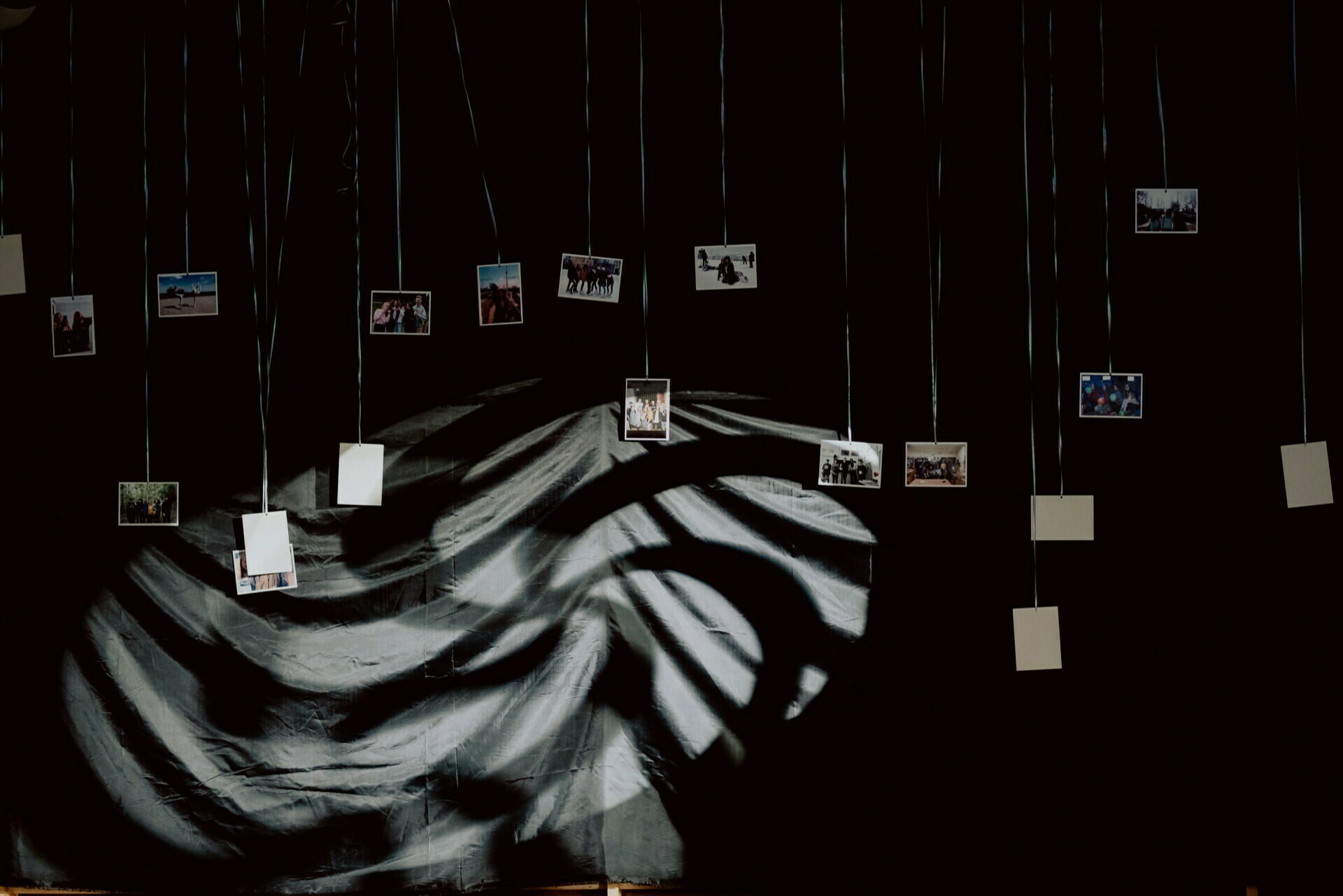 A dark room with various small photographs suspended from the ceiling on strings, presenting a stunning photo-reportage of events. The photos are scattered at different heights and angles, creating a floating effect. The background depicts a dramatic black and white shadowed fabric or texture.  