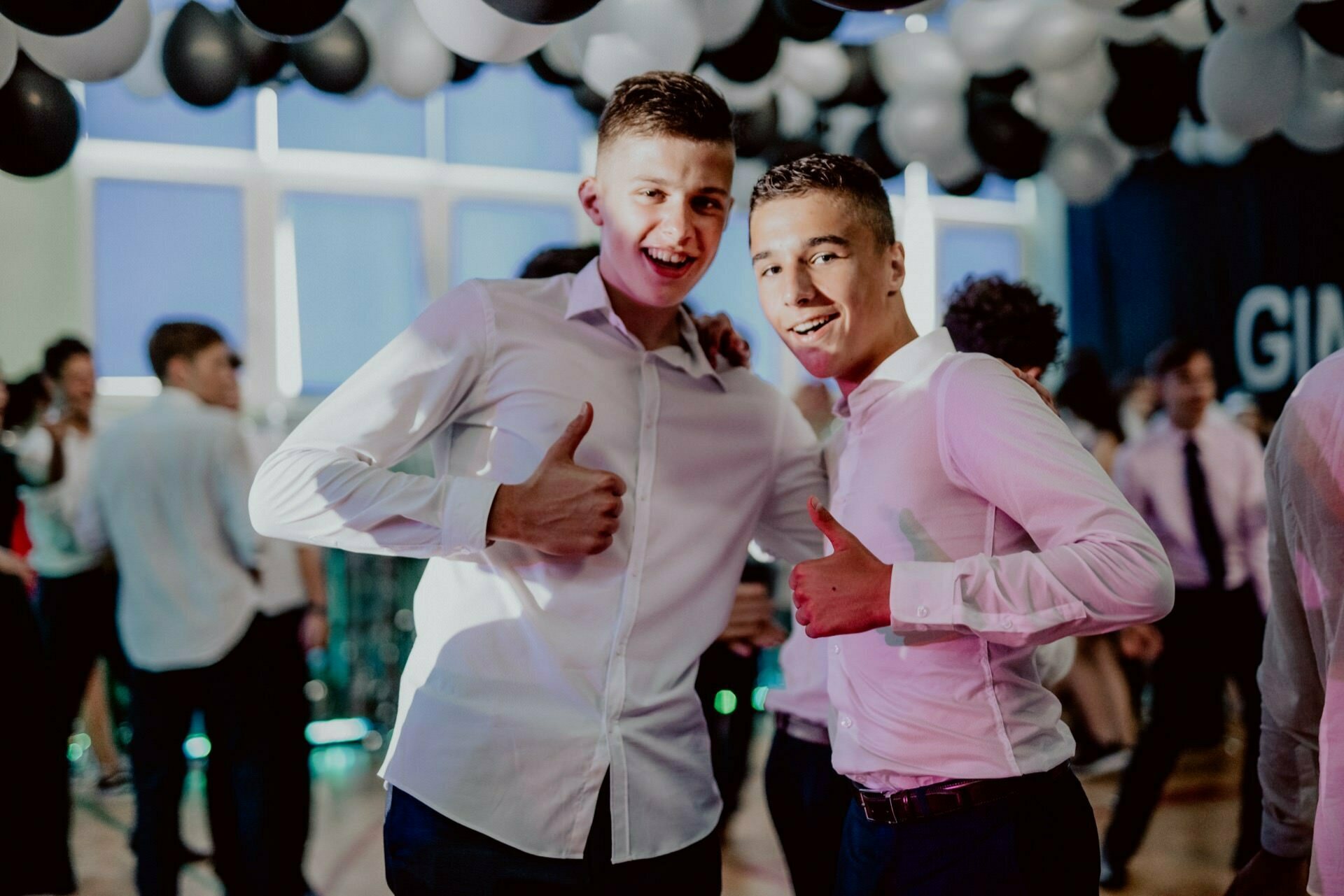 Two young men in white shirts raise their thumbs and smile at the camera during a lively event in the hall, captured as part of an event photo essay, with black and white balloons and other people in the background.