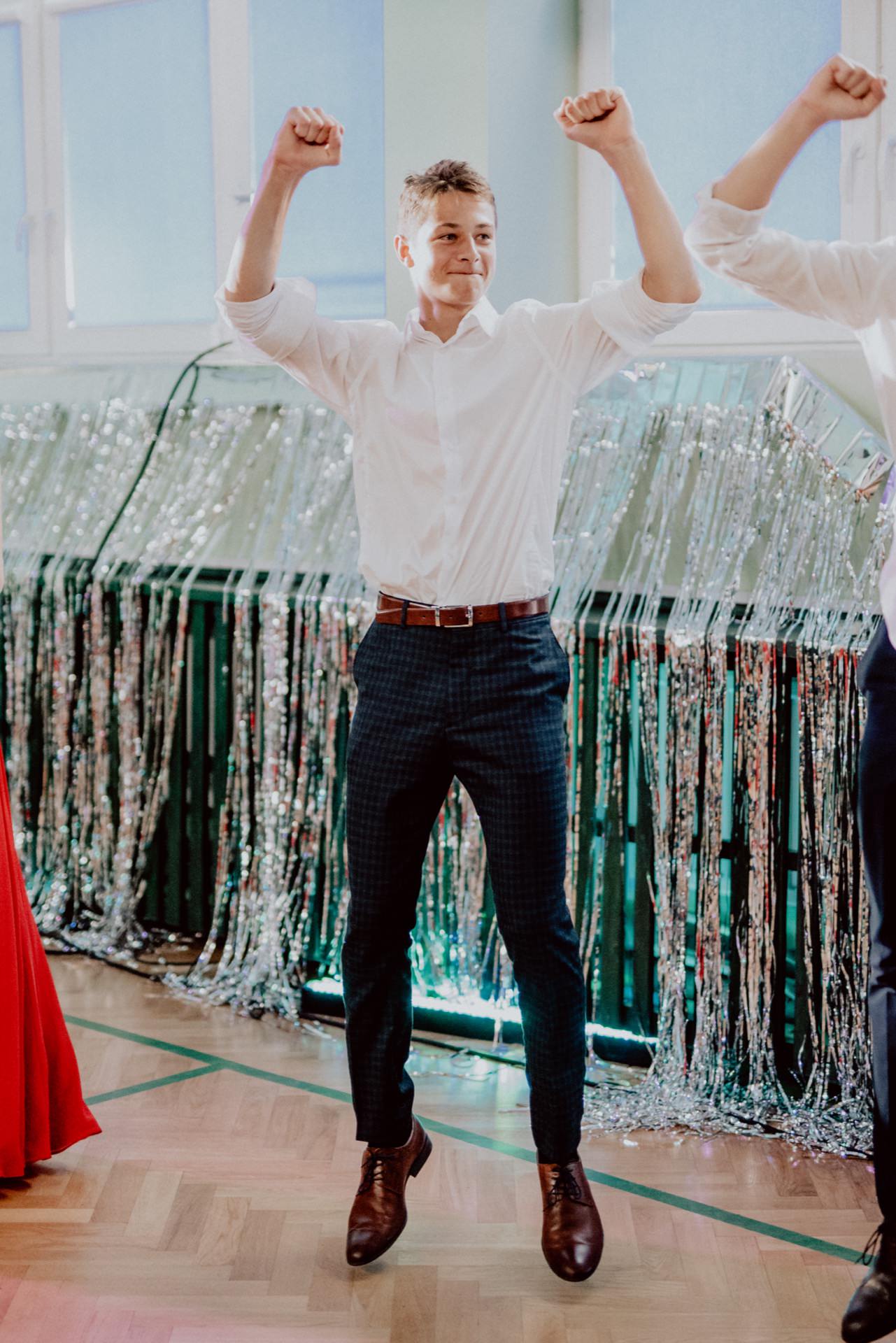 A man dressed in a white shirt and plaid pants dances with his arms raised in a room decorated with silver streamers. The bright atmosphere, enhanced by the large windows, conveys his joy as he smiles and enjoys the moment - the perfect scene for any photographic event. 