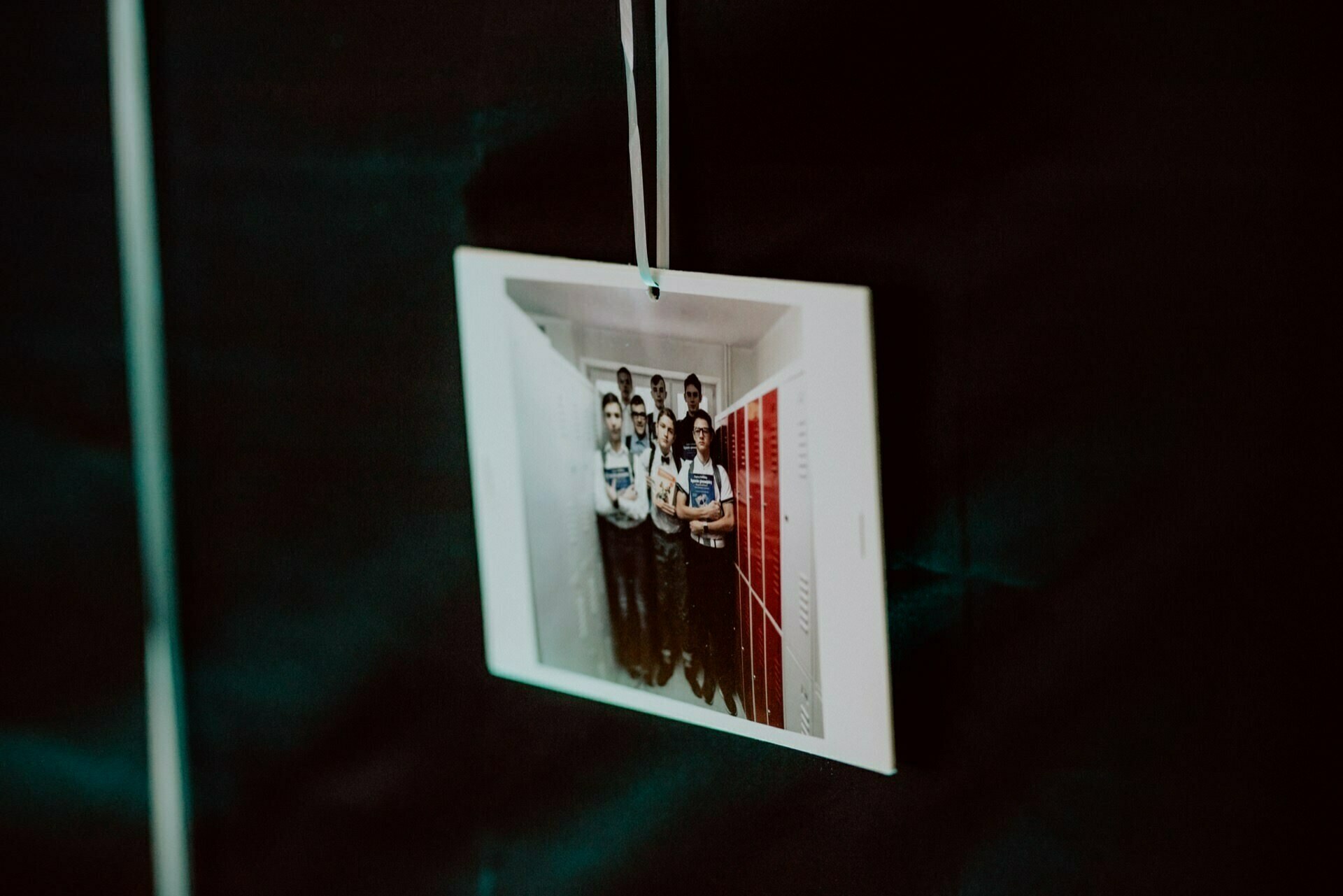 Polaroid photo hanging on black background. The Polaroid photo, taken as part of a photo report of the events, shows a group of people standing together in a corridor with red cabinets on the right. 