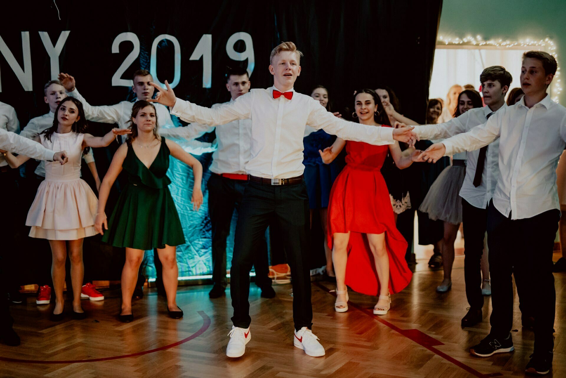 A group of young people dressed in formal attire dance energetically at a party with the year "2019" in the background. The central figure, a young man in a white shirt and red bow tie, stands with his arms outstretched, surrounded by friends - the perfect moment for event photography. 