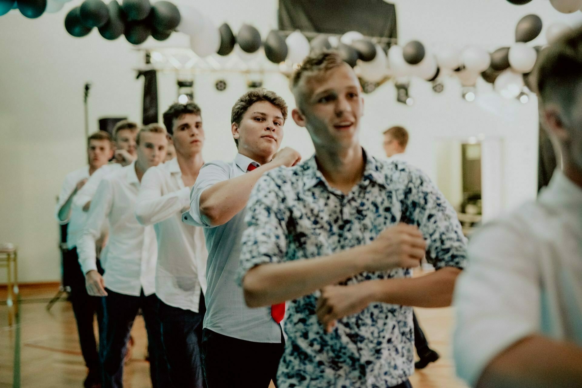 A group of young men dressed in semi-formal attire dance in a line under black and white balloon decorations. The room gives the impression of a decorated hall, and the atmosphere is lively and festive - perfect for a photo essay of events in Warsaw. 