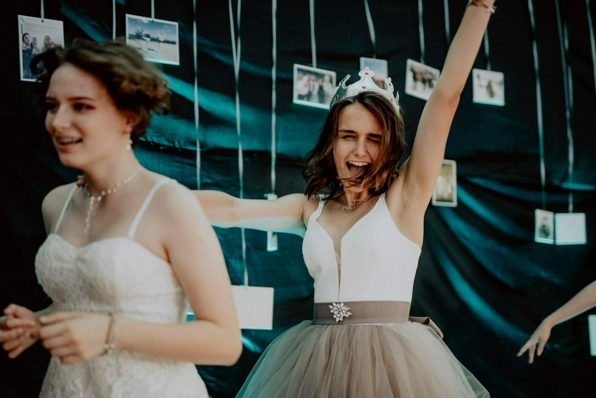 Two young women in formal dresses celebrate at an event. The woman in the foreground is wearing a white lace dress, while the woman in the back, also in a white dress with a tulle skirt, wears a crown and has her hands raised in a gesture of excitement. This vivid photo recap of the event captures their joyous moment.  