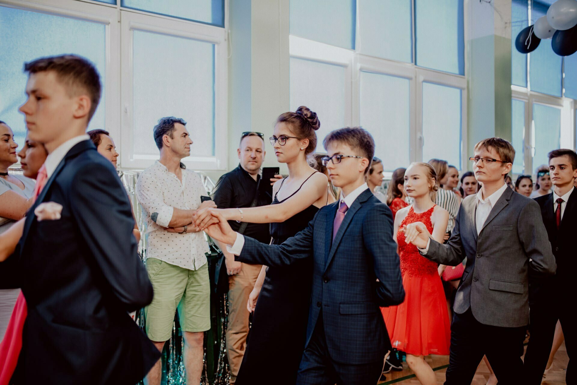 A group of young people dressed formally, some in suits and others in dresses, dance in pairs at a party held indoors. The dancers are watched by spectators, including a man in a white shirt and green shorts. The room is bright, with large windows and decorations - perfect for event photography by event photographer Warsaw.  