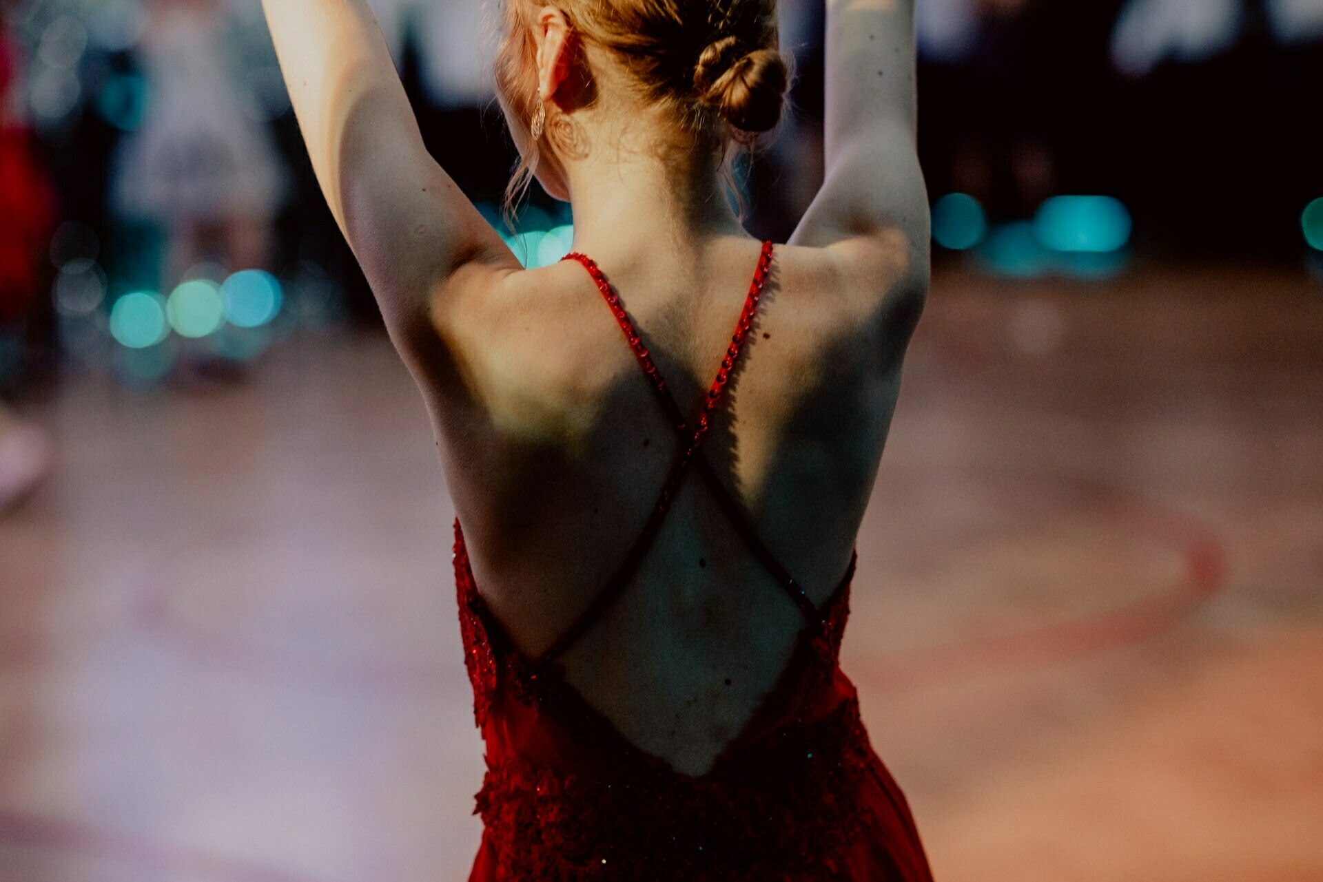 A person with light brown hair pinned up in a bun stands with their hands raised, face turned away from the camera. They are wearing a red backless dress with thin straps crossing in an X. The background is blurred, showing a dimly lit room with a few people and lights - perfect for event photography at an exclusive event. 