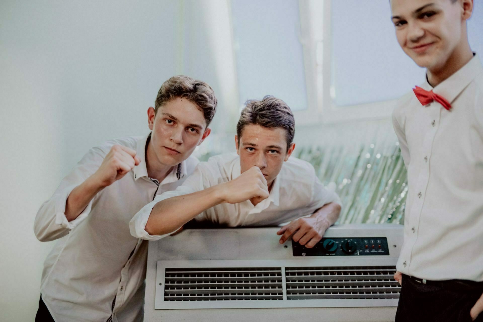 Three young men in white shirts pose for a photo. The two on the left show a playful strongman gesture with their fists, while the one on the right smiles while wearing a red bow tie. This excellent photo is an example of event photography near a large white machine with control knobs and vents.  