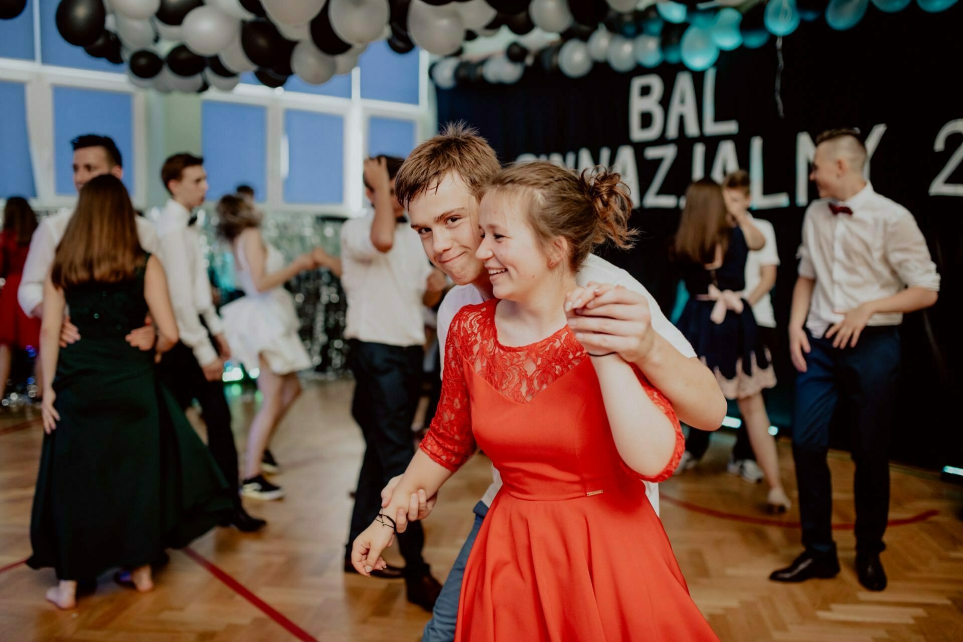 A young couple, a girl in a red dress and a boy in a white shirt, dance together and smile. Other people dance in the background under black and white balloons during a lively prom. "BAL" is visible among the decorations, perfectly captured for the event photo report by event photographer Warsaw.  