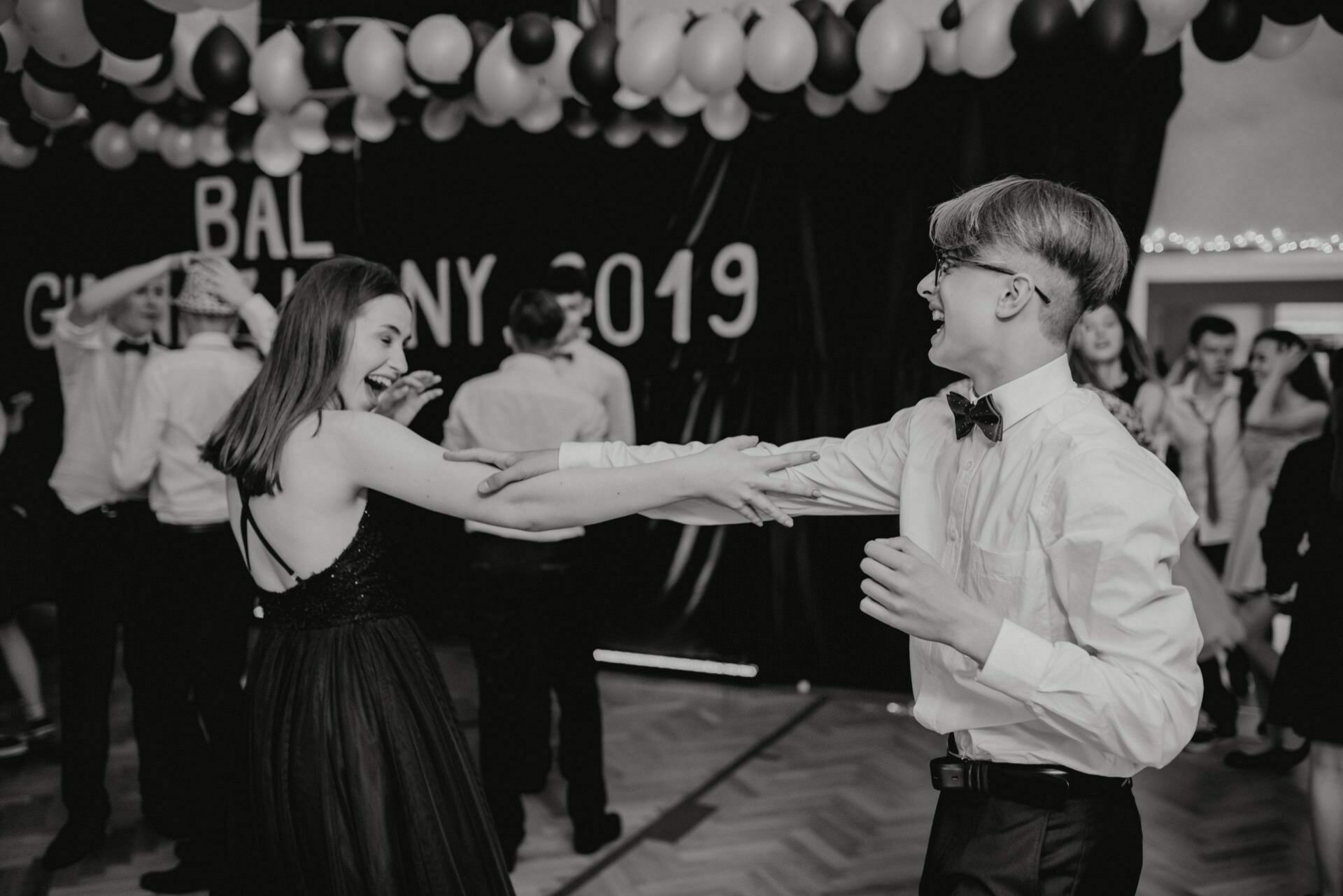 In the black and white event photo, a young couple dressed formally smiles and holds hands as they dance in a decorated room with balloons overhead and "2019 Gymnasium Ball" in the background. Other participants are visible in the background. (Event photo warsaw)  