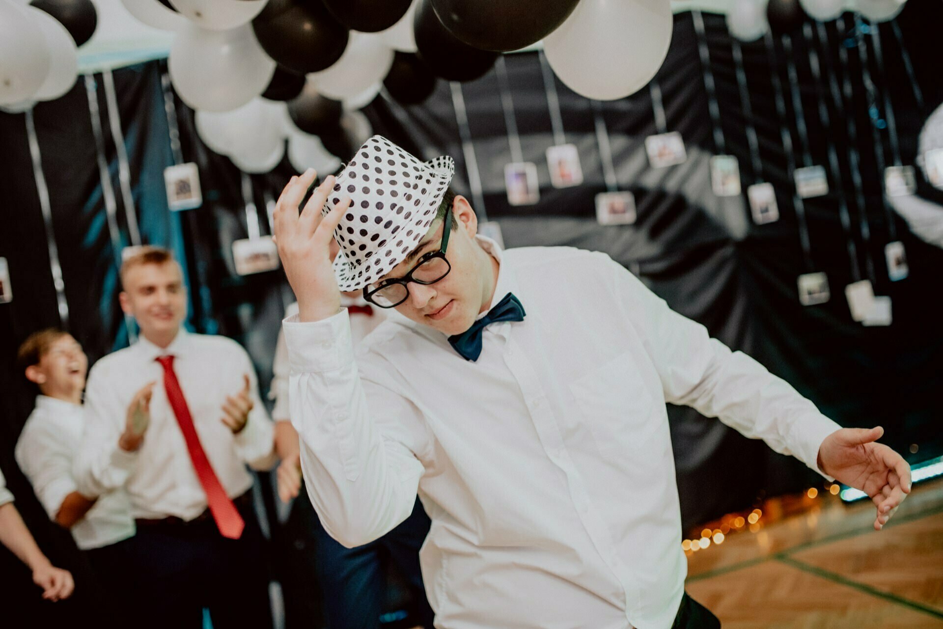 A person dressed in a white shirt, black bow tie, glasses and a pea hat tilts his hat while dancing at an elegant party. Others, dressed formally, clap in the background. The ceiling is decorated with black and white balloons and hanging photographs - a perfect scene for event photography.  