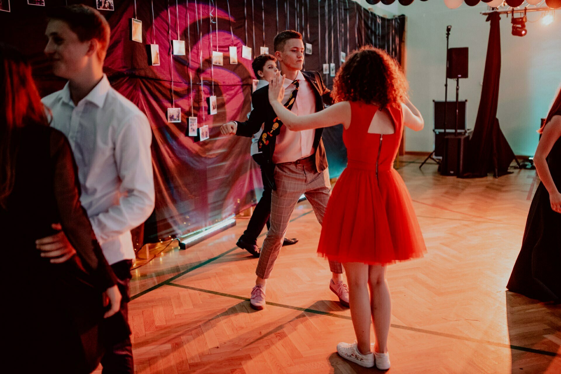 People are dancing in a dimly lit room decorated with a dark background and hanging pictures. Two people in the foreground are dancing, one dressed in a red dress and the other in a jacket and patterned pants. The other dancers are visible in the background, capturing the essence of event photography.  