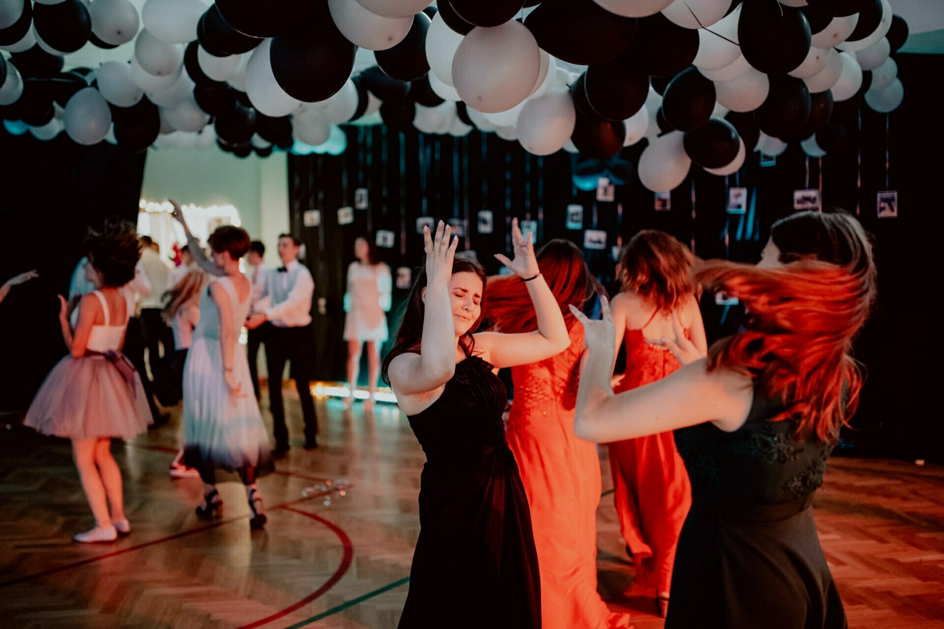 People in formal attire enthusiastically dance under a ceiling decorated with black and white balloons. A few people in the background also take part in the festivities taking place in a dimly lit venue, which was perfectly captured by event photography professionals. 