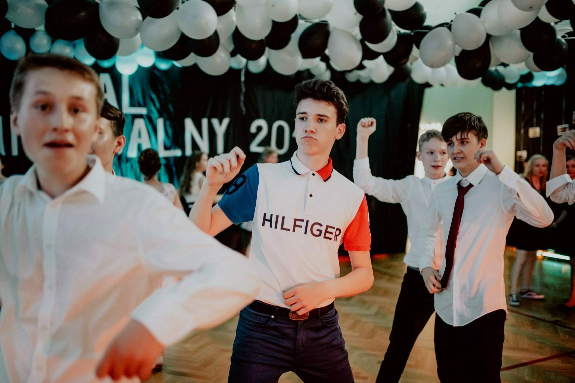 A group of young boys dressed in formal and casual attire dance under a ceiling covered with black and white balloons. One of the boys in the foreground is wearing a white polo shirt with "Hilfiger" written on it. Perfectly captured by the event photographer warsaw, in the background is a banner with partially visible text.  