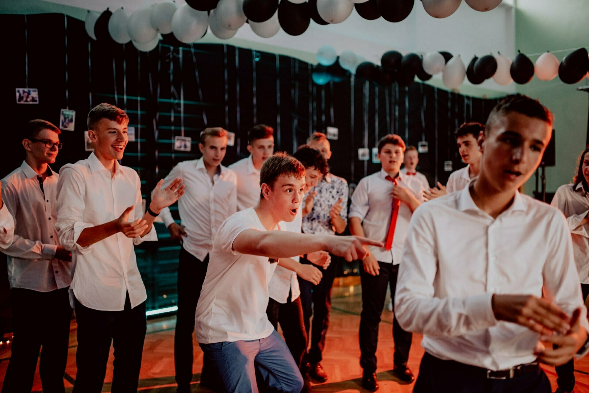 A group of young men, dressed mostly in white shirts, dance and play in a dimly lit room with black and white balloons over their heads. Some are gesticulating enthusiastically, while others are looking on and smiling, creating a lively and joyful atmosphere, perfect for a photographic event. 