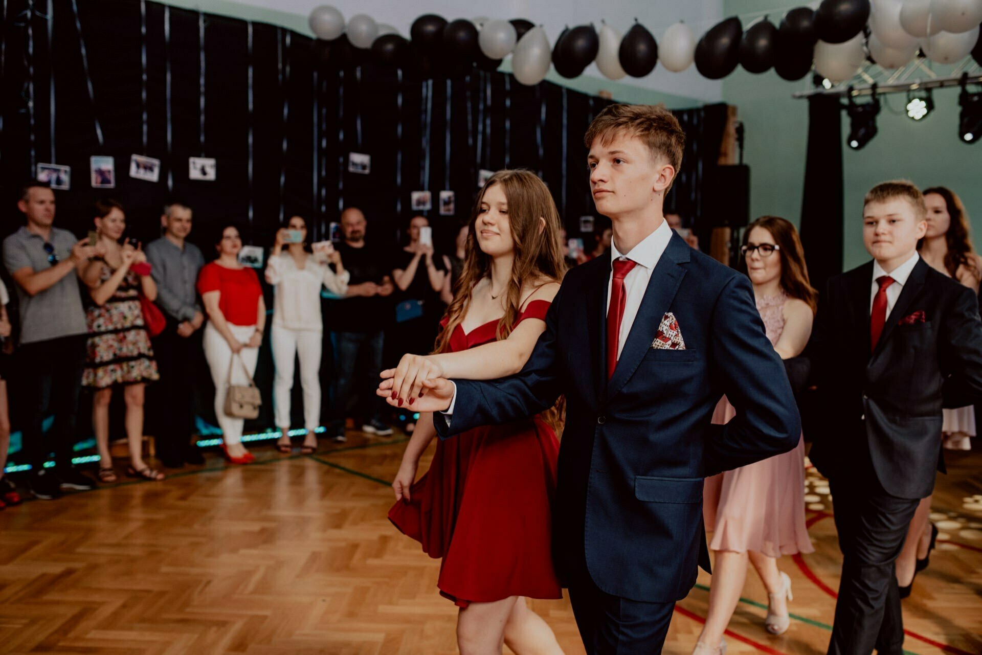 A young couple in formal attire dances in the middle of a hall decorated with black and white balloons, while the crowd watches and takes pictures. The young man is dressed in a dark suit, while the young woman dazzles in a red gown, which was beautifully captured by an event photographer Warsaw. 