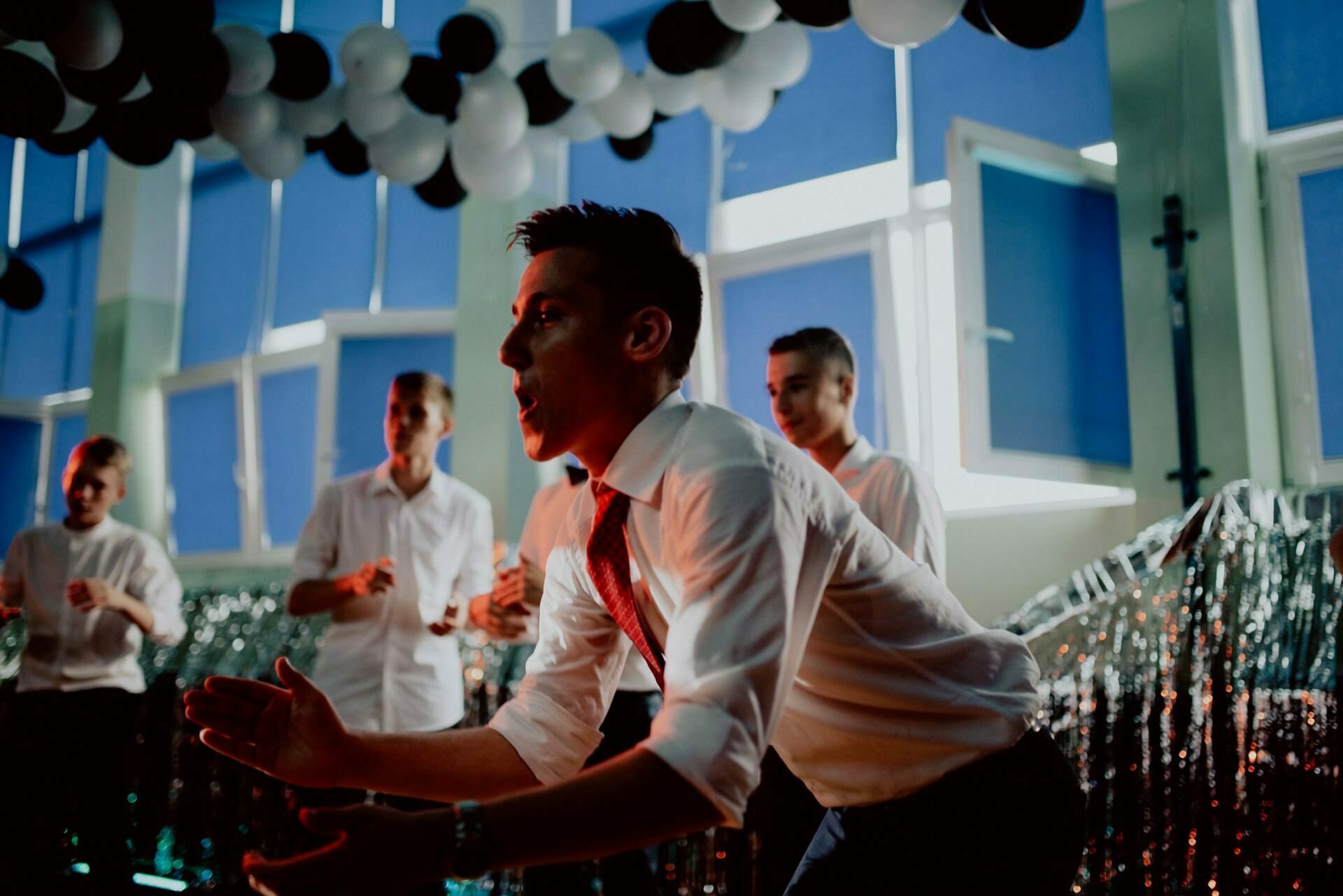 A group of people are at a party decorated with black and white balloons and glittering garlands. One person in the foreground in a white shirt and red tie appears to be clapping or cheering. Others in the background are also enjoying the event, creating the perfect scene for an event photographer.  