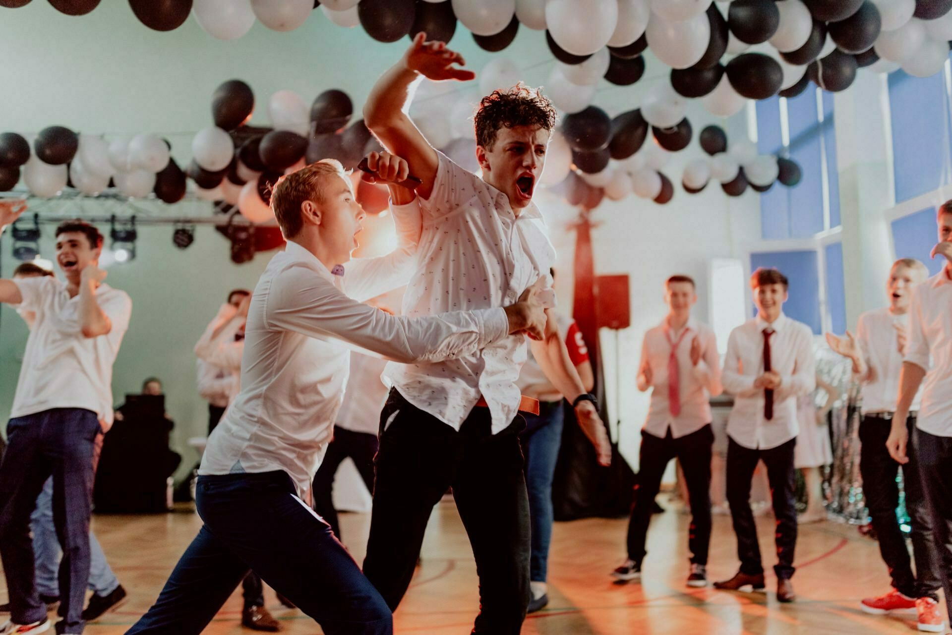 A lively group of young adults dancing energetically at a party. The room is decorated with black and white balloons, and sunlight streams in through the large windows. The two men in the lead are passionately dancing and singing, dressed in white shirts and dark pants - a perfect scene for event photography.  