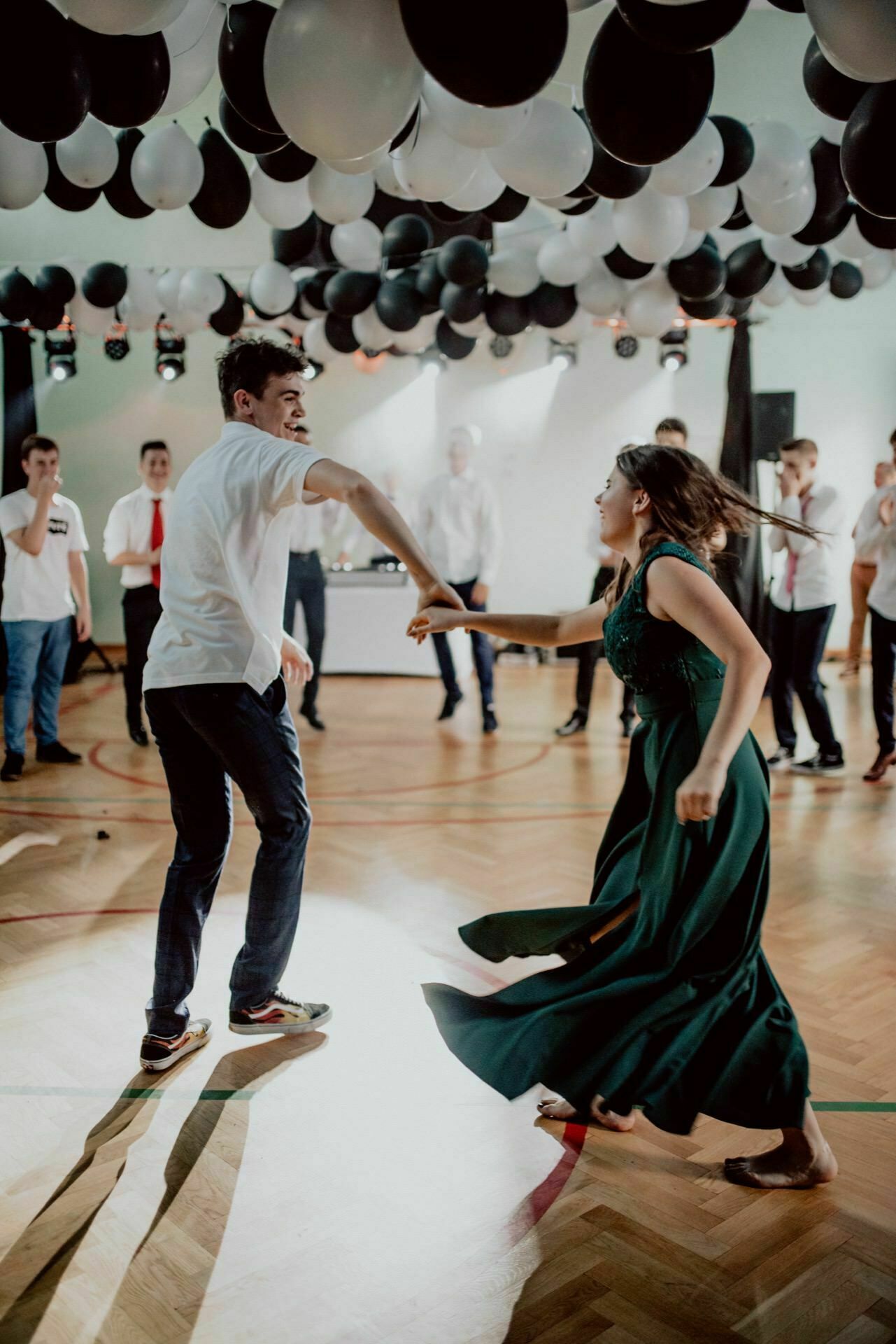 A young man and woman dance energetically under black and white balloons in a decorated room with a wooden floor, while other people stand in the background and watch. The woman is dressed in a green dress and the man in a casual white shirt and dark pants - a perfect shot for event photography. 