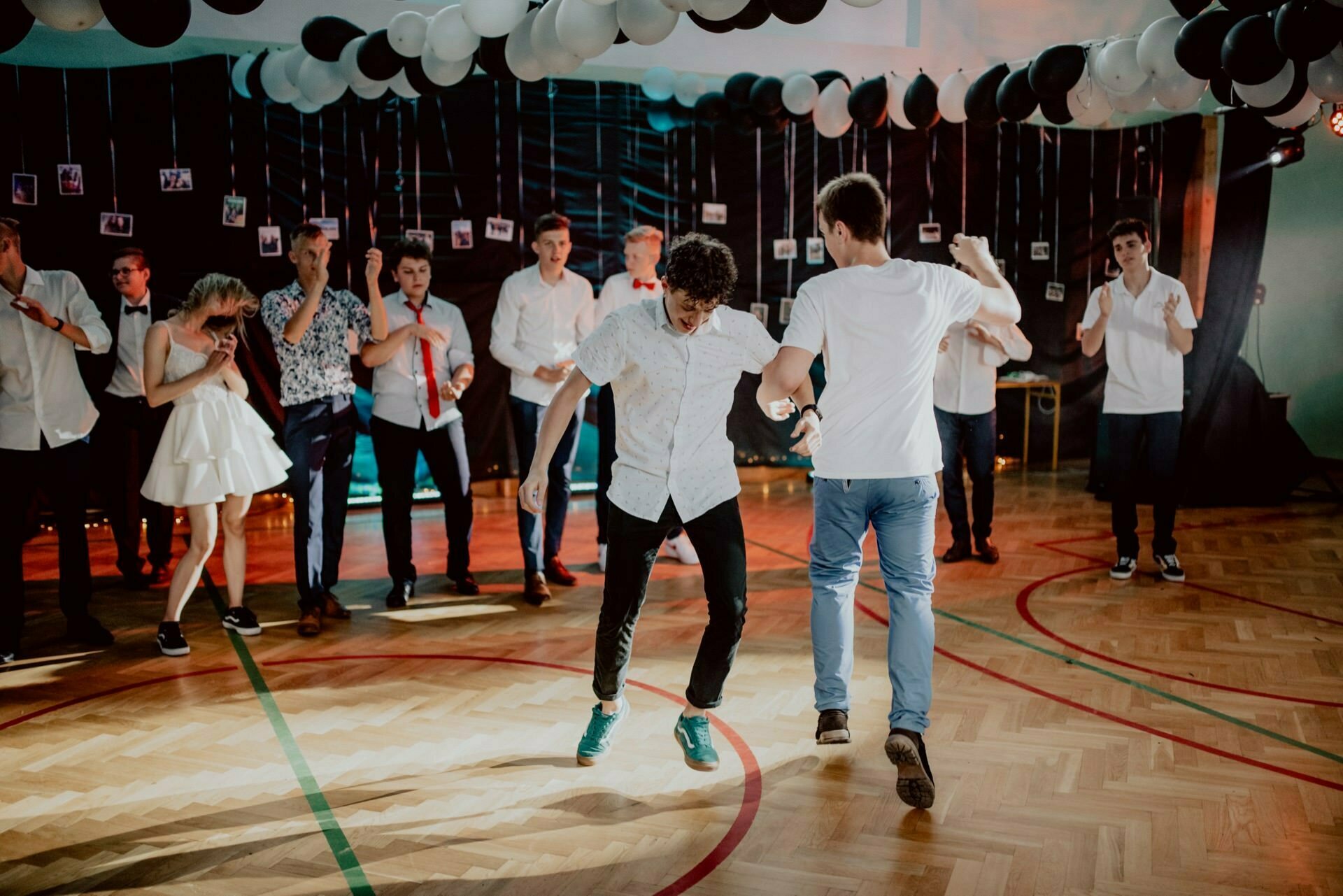 A vibrant party scene features two people dancing energetically in the middle of a gymnasium that has been turned into a party, decorated with black and white balloons and hanging photos. Spectators dressed in semi-formal attire stand around, watching and cheering, creating the perfect setting for photo coverage of the events. 