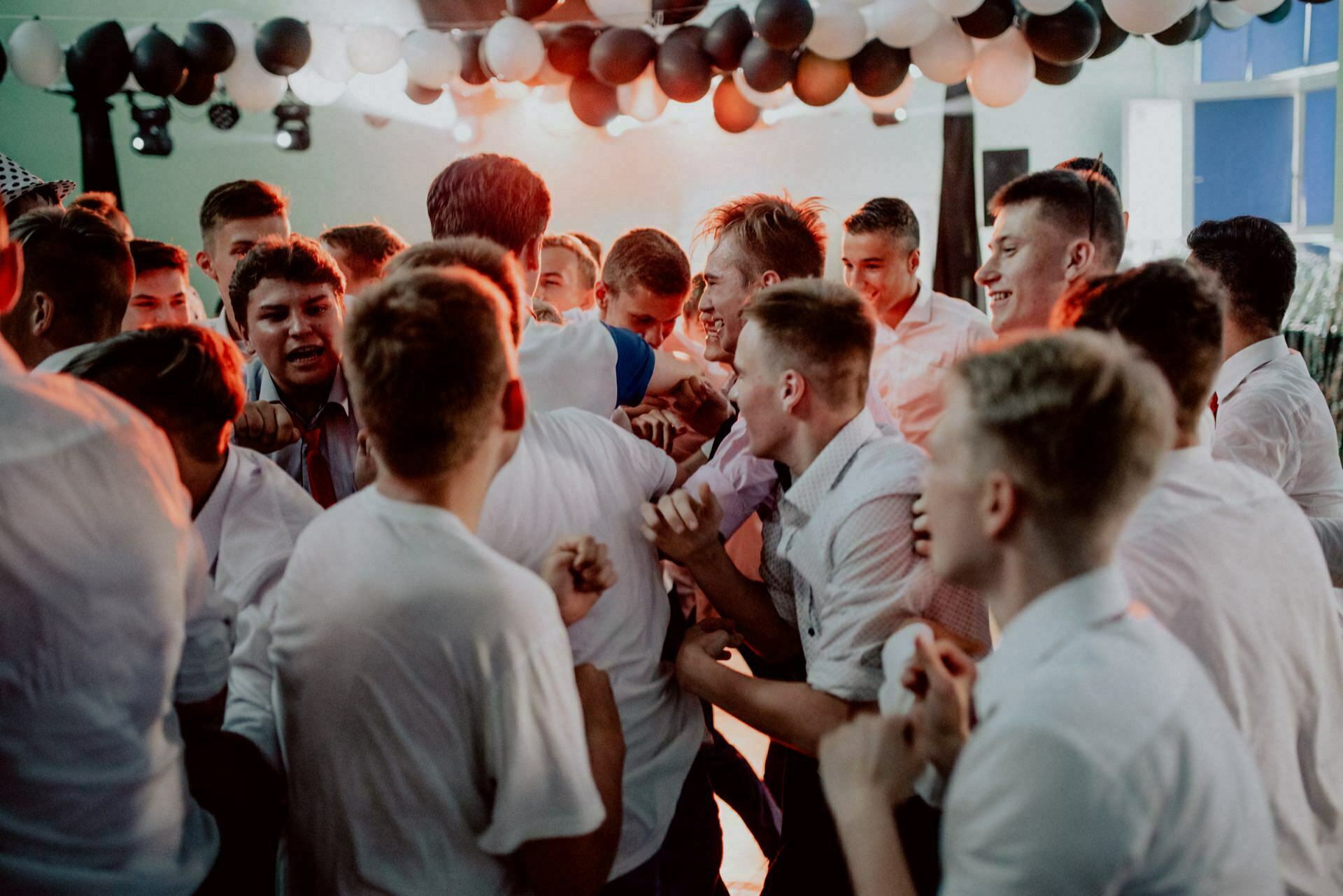 A group of young men in white shirts stand close together under black and white balloons during what appears to be an event or party in the room. Some are cheering or laughing, creating a lively and energetic atmosphere, which is perfectly captured by the event photography. 