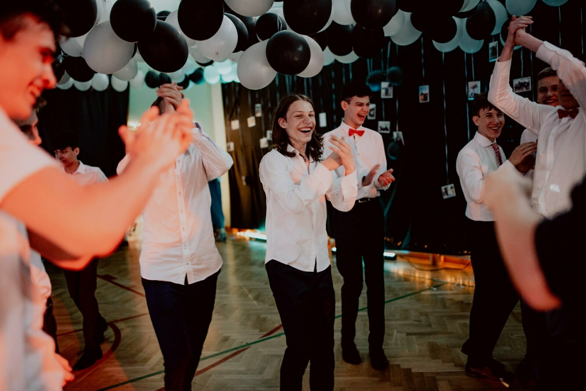 A group of young people dressed in formal attire dance and clap under black and white balloons during a festive event. They look jubilant and are in a decorated indoor space with black curtains and garlands of light, perfect for capturing event photo coverage by any event photographer Warsaw has to offer. 