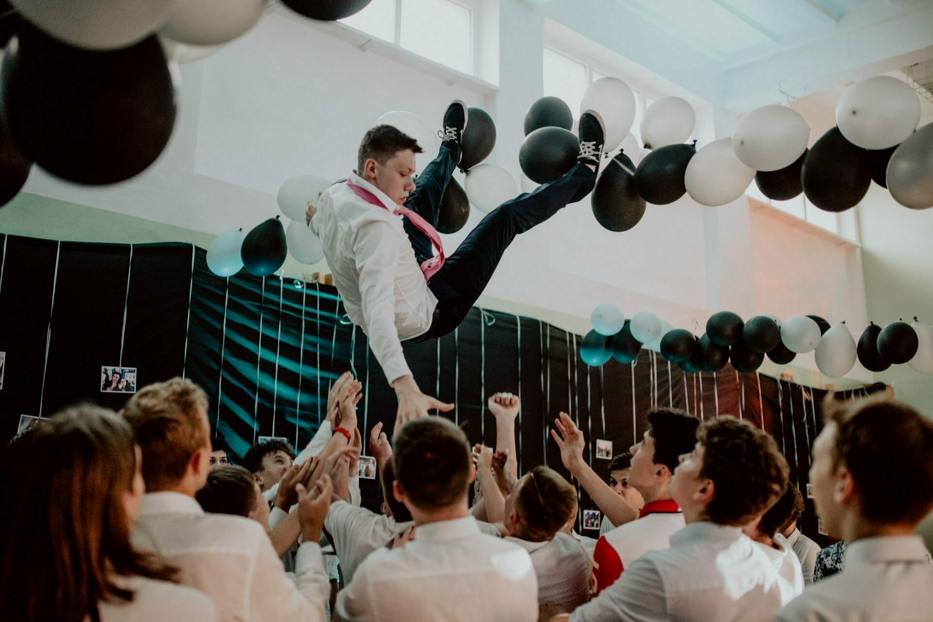 A group of people lifts a person in formal attire into the air during a ceremony held in a room. The room decorated with black and white balloons is filled with the joyful cheers of people in white shirts. This vibrant scene captures the essence of event photography done by a skilled event photographer warsaw.  