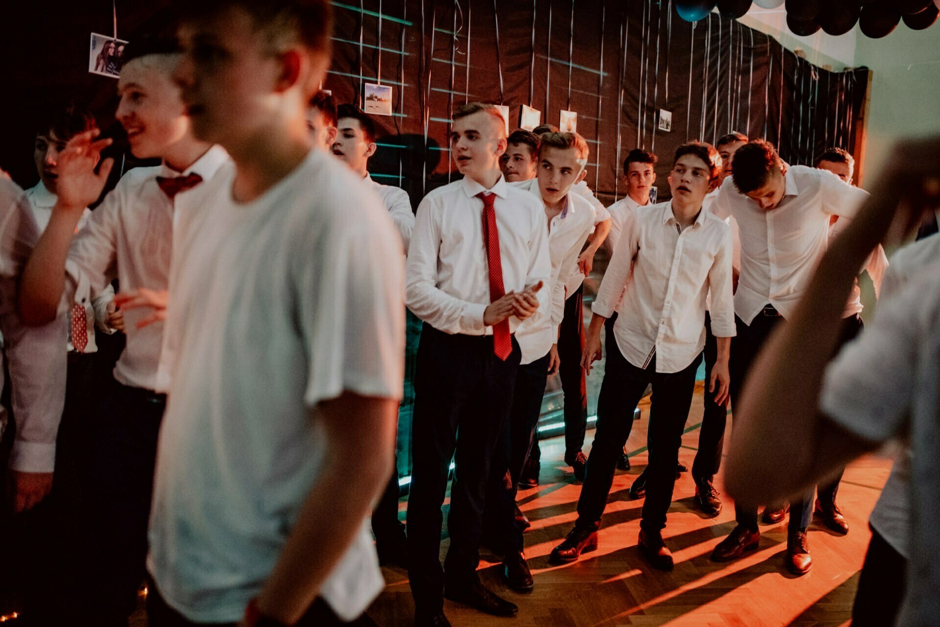 A group of young men, some in white shirts and red ties, others in plain white T-shirts, gather in a dimly lit room with a tiled wall decorated with pictures. The scene seems animated, capturing the essence of the photo-reportage of events, as some look toward the camera and others engage in conversation. 