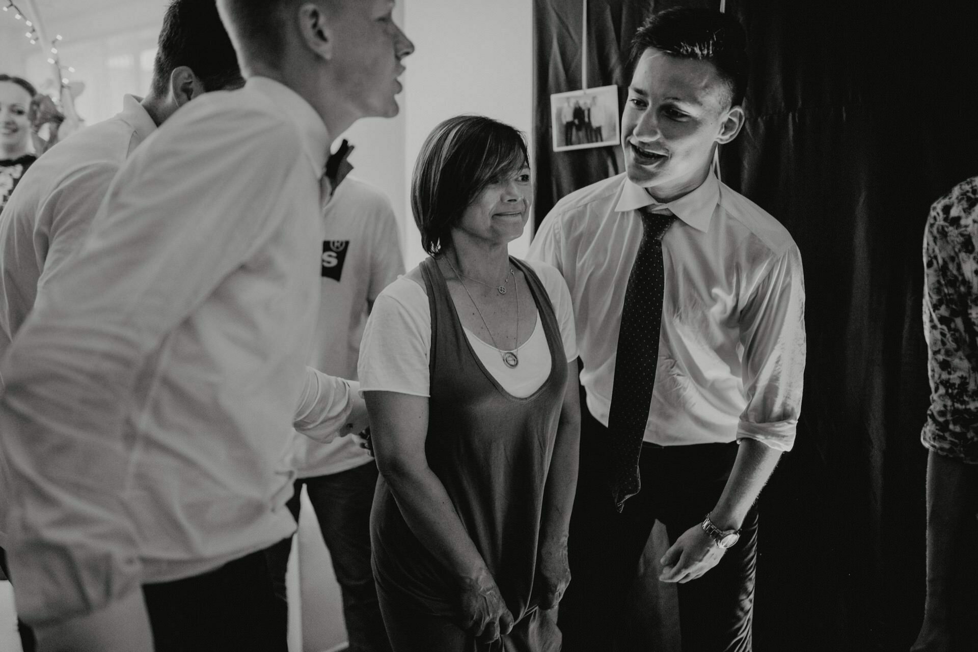 Black and white event photo showing people at a social event. The woman in the middle is smiling, wearing a casual top with a necklace. On either side of her stand two men dressed in shirts and ties, carrying on a conversation and smiling. More people can be seen in the background, capturing the essence of the photo event.   