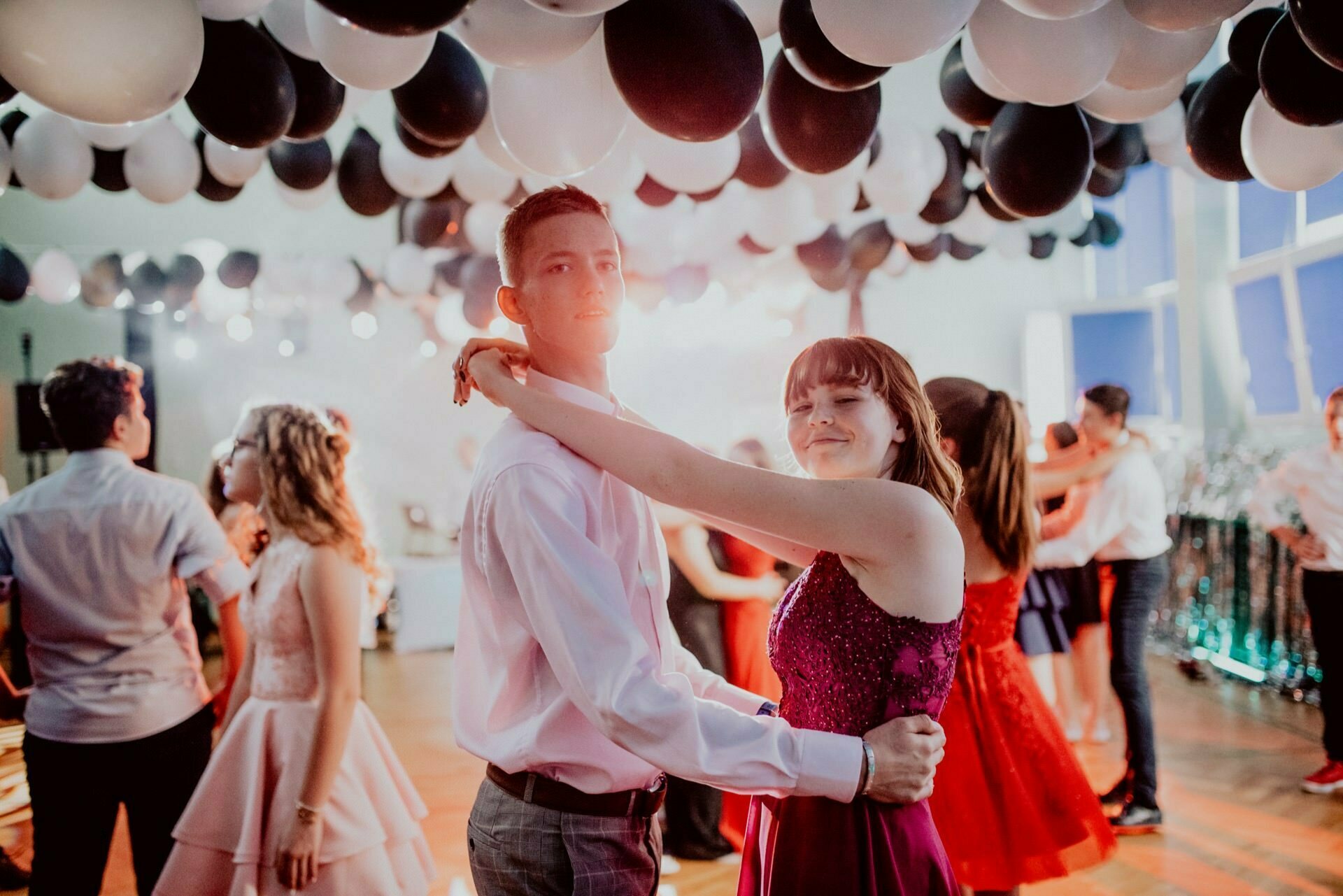 A young couple is dancing under a ceiling filled with black and white balloons. The girl is wearing a red sparkly dress and the boy is wearing a white shirt with a tie. There are also other couples dancing in the background. Soft lighting creates a warm, festive atmosphere, perfect for capturing moments during event photography.   