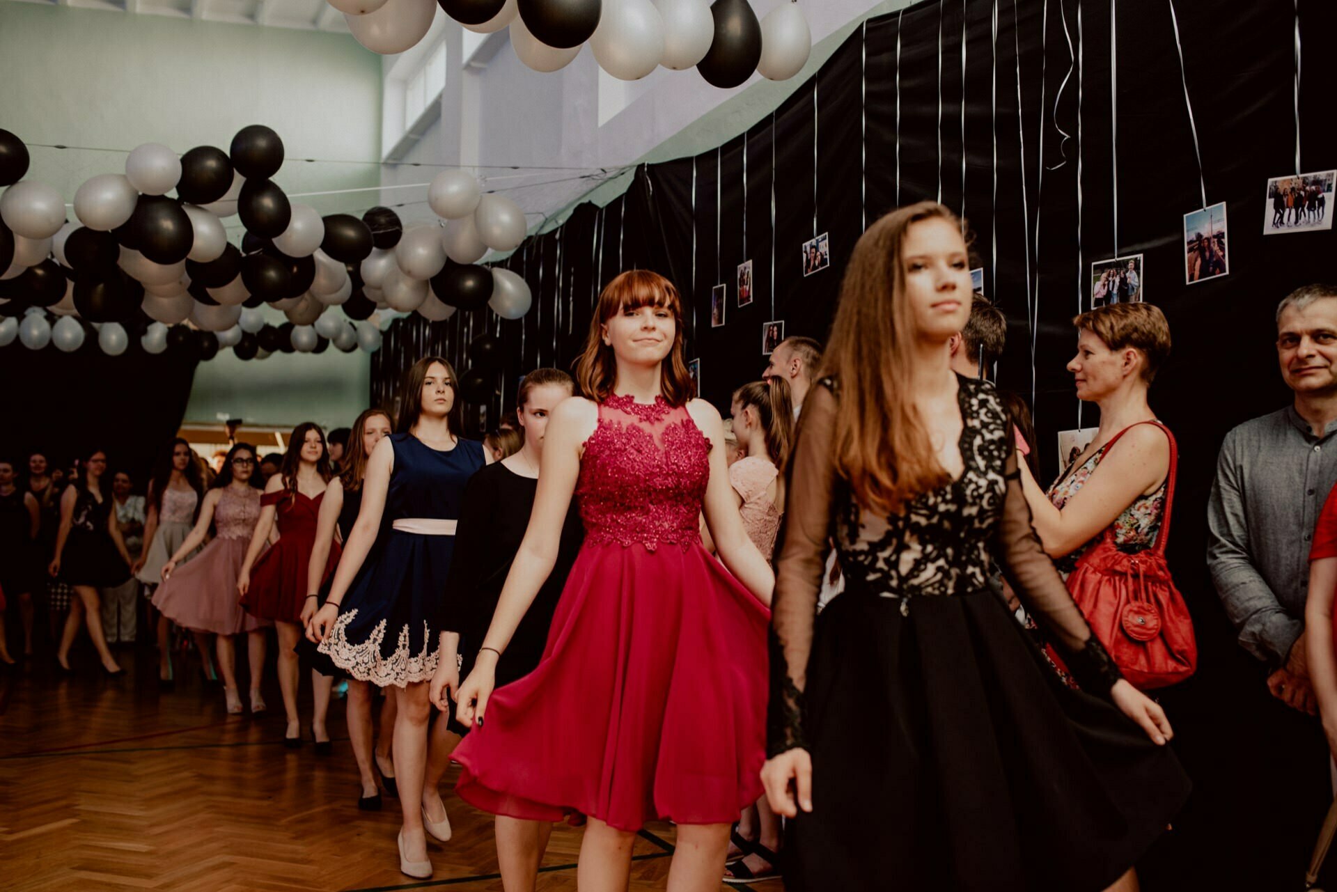 A group of people dressed in formal attire stand in line during a ballroom dance. The set design includes black and white balloons arranged in arches overhead and a wall decorated with strings of photos. The group is led by a woman in a red dress and another in a black dress - perfect for event photography.  