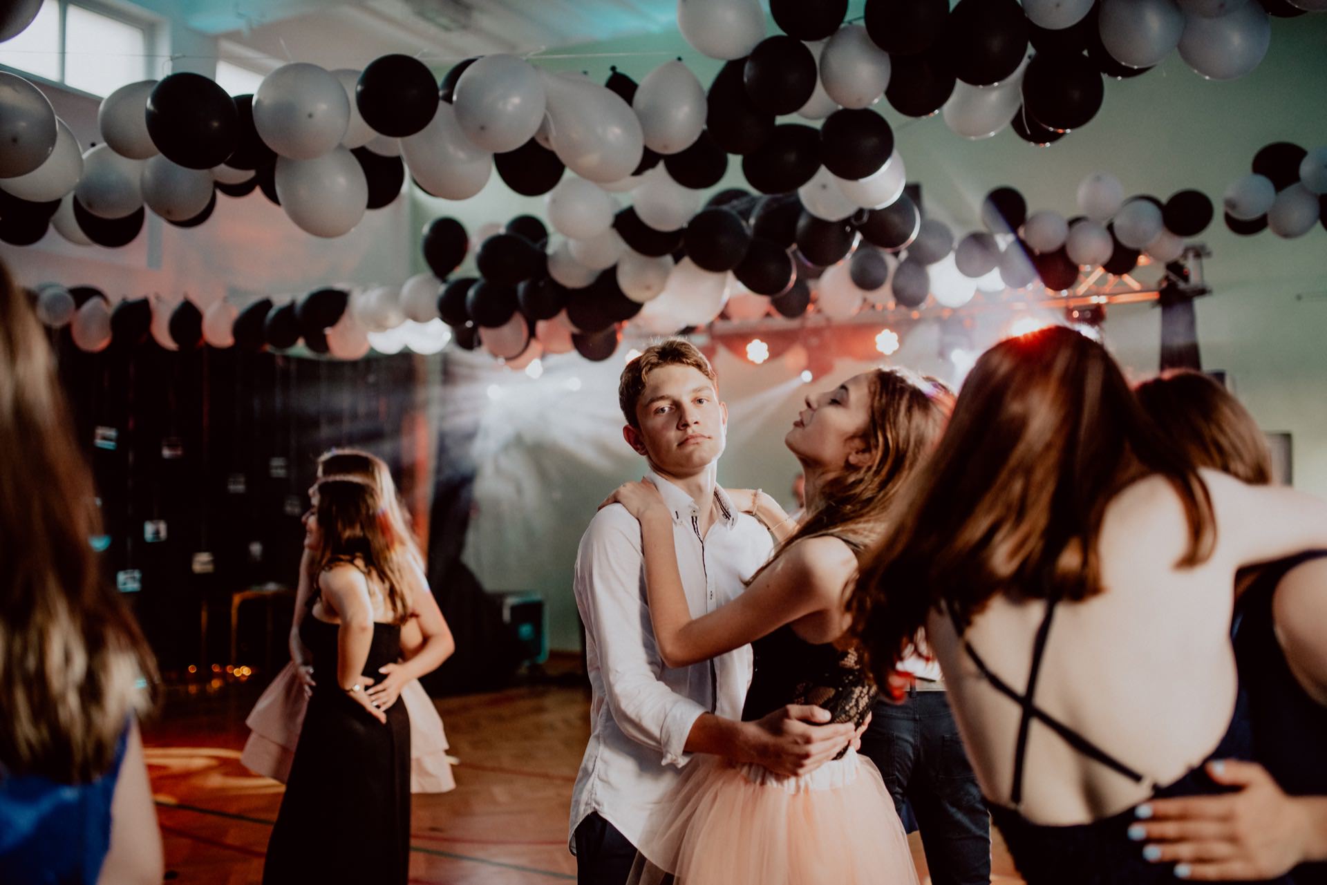 In this captivating photo report of the event, a man and woman couple dance under a ceiling decorated with black and white balloons. The man looks into the camera, his expression neutral, while the woman rests her head on his shoulder. Other couples also move gracefully in the dimly lit room.  