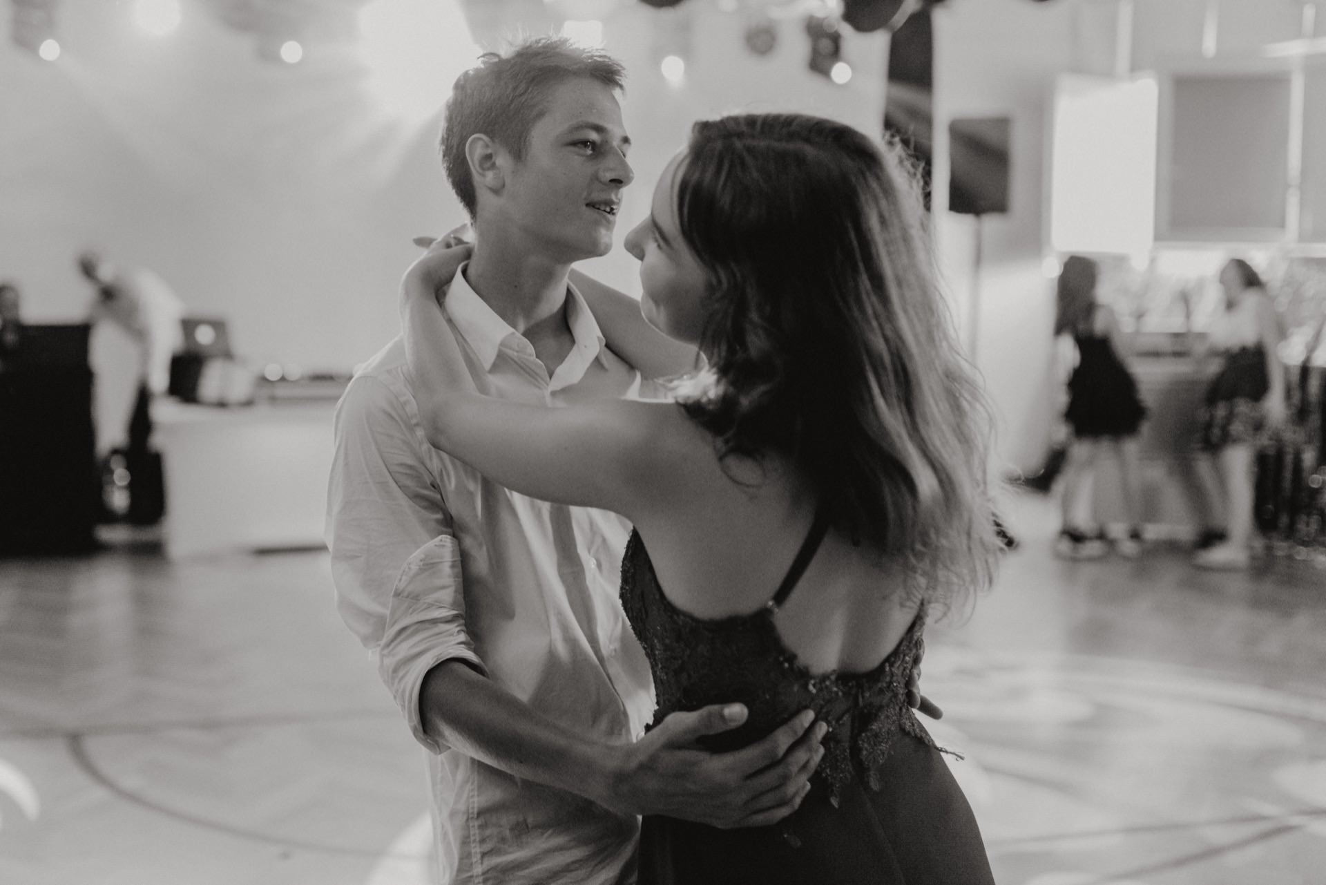 Black and white photo of a young couple dancing closely at a party or event. The man, dressed in a light-colored shirt, looks at the woman embracing him. In the background you can see other people, a DJ booth and a lit dance floor - capturing the essence of event photography.  