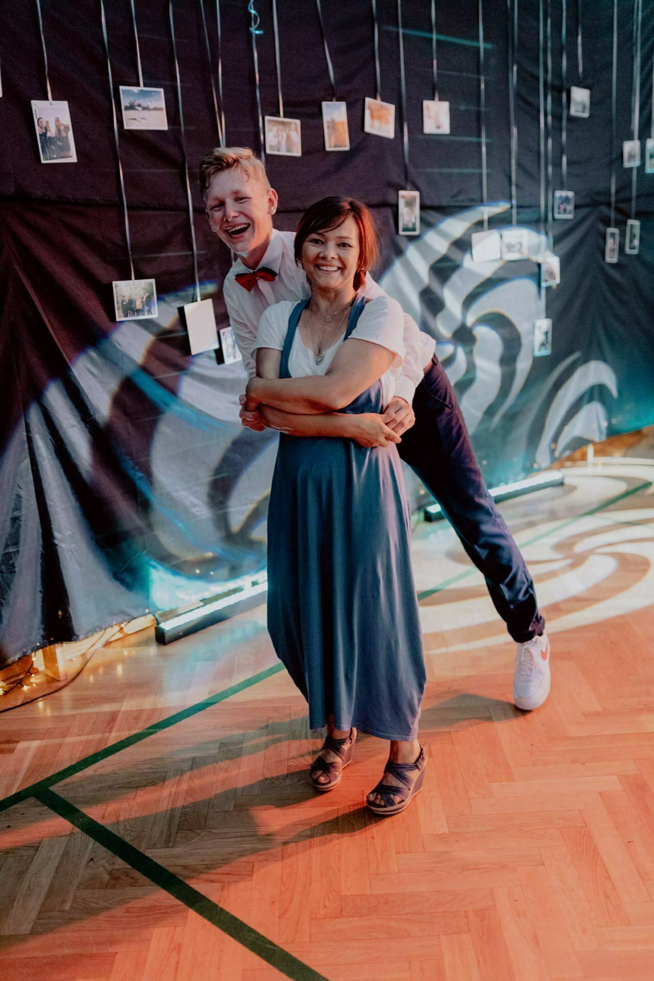 A man and woman pose happily together in a room, with the man standing behind and next to the woman, leaning forward. The woman is wearing a blue dress and crossed arms, while the man is dressed in a white shirt and bow tie. There are several hanging photographs and decorative lighting in the background, perfectly capturing this event photography.  