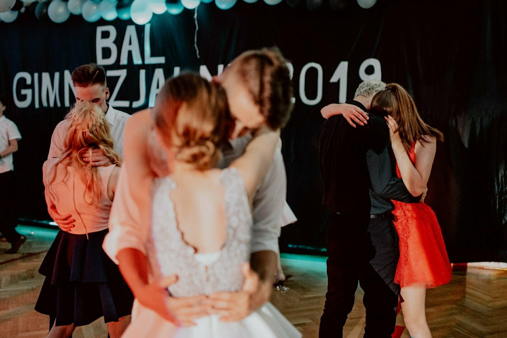 Young couples slowly dance at a covered party with balloons and a black banner on the wall that reads "Gymnasium Ball 2019." The floor is wooden, everyone is dressed in formal attire, and an event photographer Warsaw captures every touching moment. 