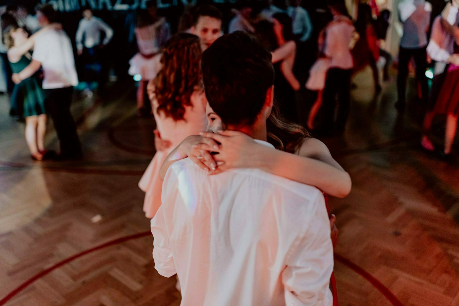 A couple dances closely during a dimly lit event, beautifully captured by an event photography specialist. The woman embraces the man by the neck while he stands with his back to the camera. Other people are dancing in the background on the wooden floor.  