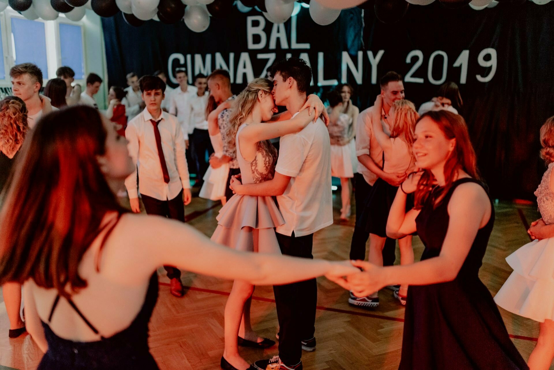 A group of young people are dancing in a decorated hall with the words "Middle School Ball 2019" written on the wall. They are wearing semi-formal outfits. The focus is on two couples dancing closely in the foreground, while others mingle and dance in the background, which was perfectly captured by event photographer Warsaw.  