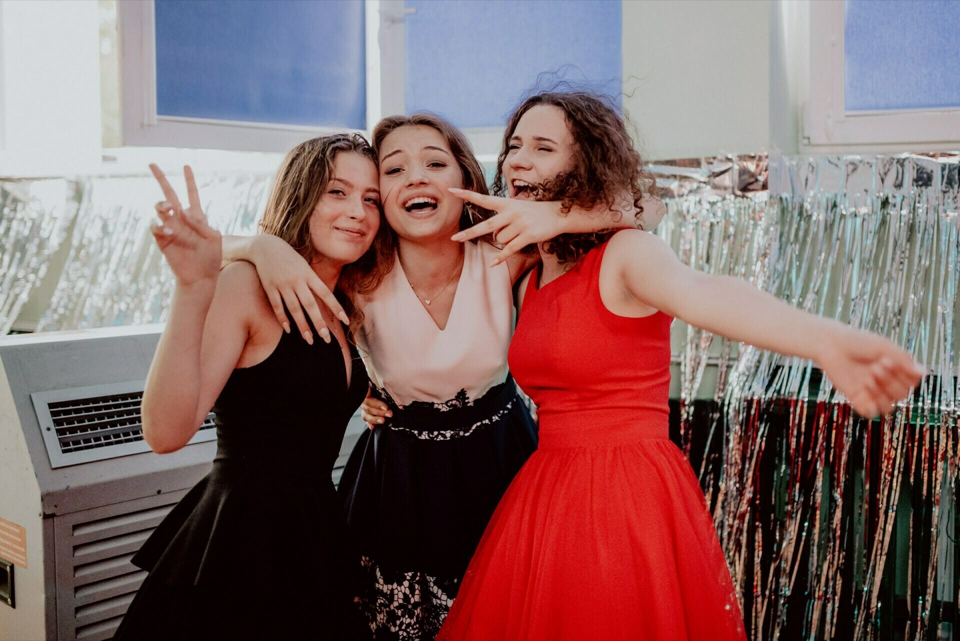 Three young women joyfully pose together. One makes a peace sign, another smiles broadly while embracing a friend, and the third extends her hand. They wear elegant gowns in black, white and red against a background of festive silver streamers - perfect for event photography by event photographer Warsaw.  