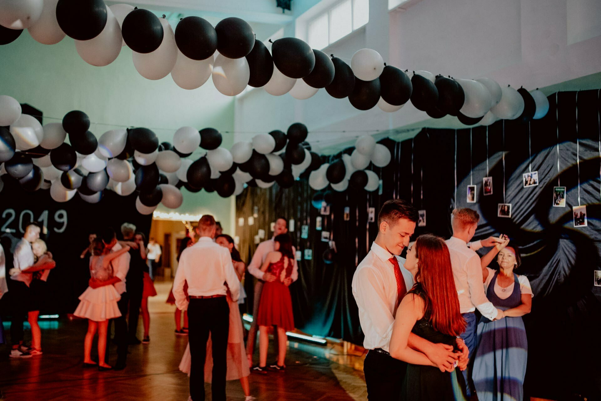 A group of people dance in a decorated room with black and white balloons hanging from the ceiling. The event appears to be formal, with the participants dressed in elegant attire. Black curtains and wall murals adorn the walls, showing the essence of the celebration through a photo report of the events by an event photographer Warsaw. The atmosphere is lively and festive.   