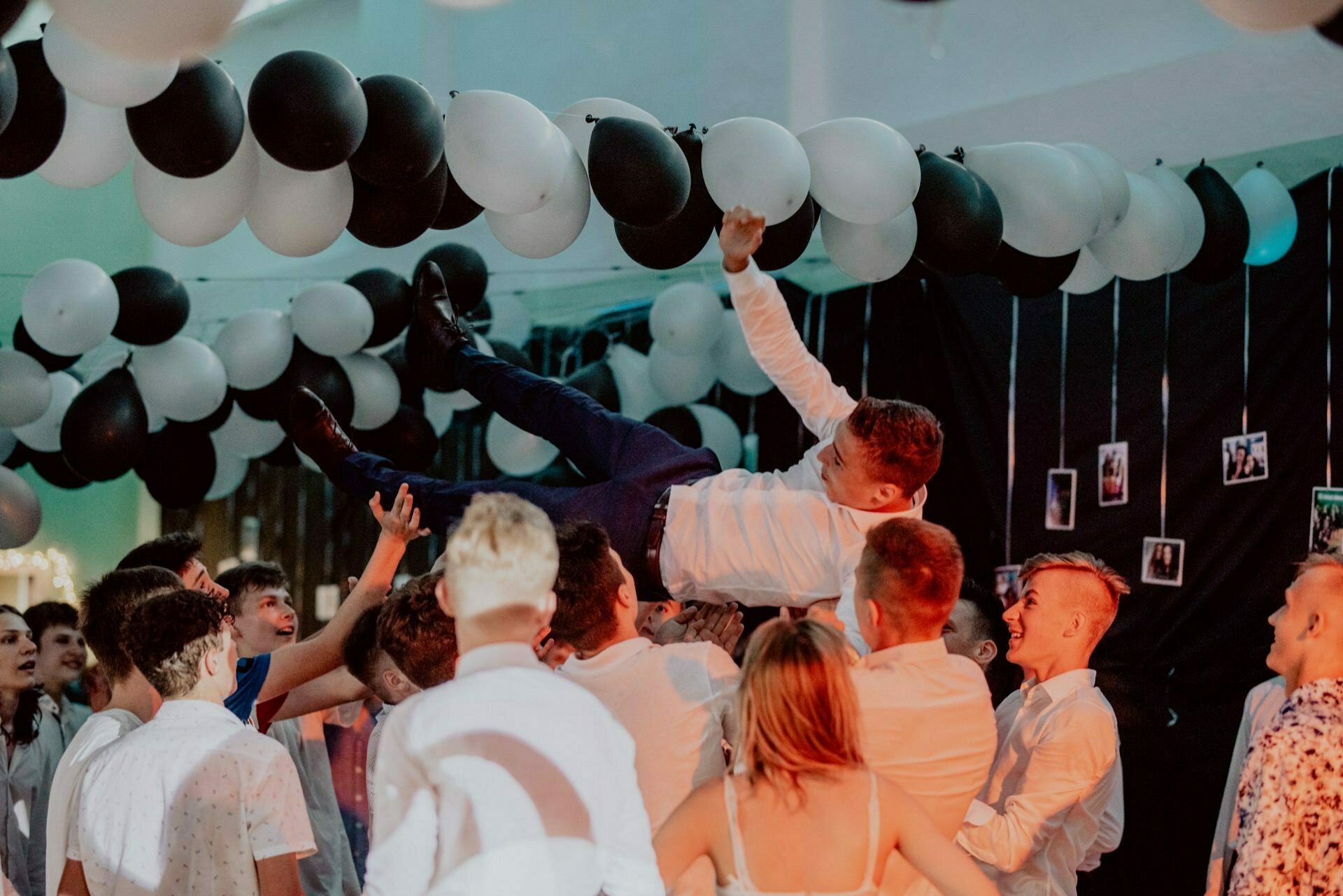 A group of people lifts a smiling man in a white shirt and dark pants into the air during a lively ceremony. Black and white balloons hang above their heads, and the photos are projected onto a black background. The crowd, which brought a festive atmosphere with its bright attire, perfectly captures the essence of event photography.  