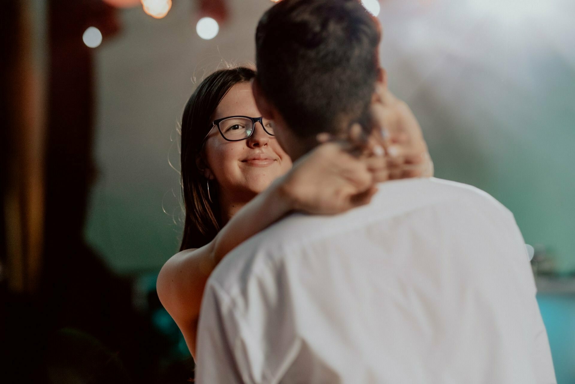 A woman with long dark hair and glasses smiles as she embraces a man from behind and hugs him around the neck. The man, dressed in a white shirt, stands with his back to the camera. The background is blurred with warm and cool lights, capturing an intimate moment typical of event photography by event photographer Warsaw.  