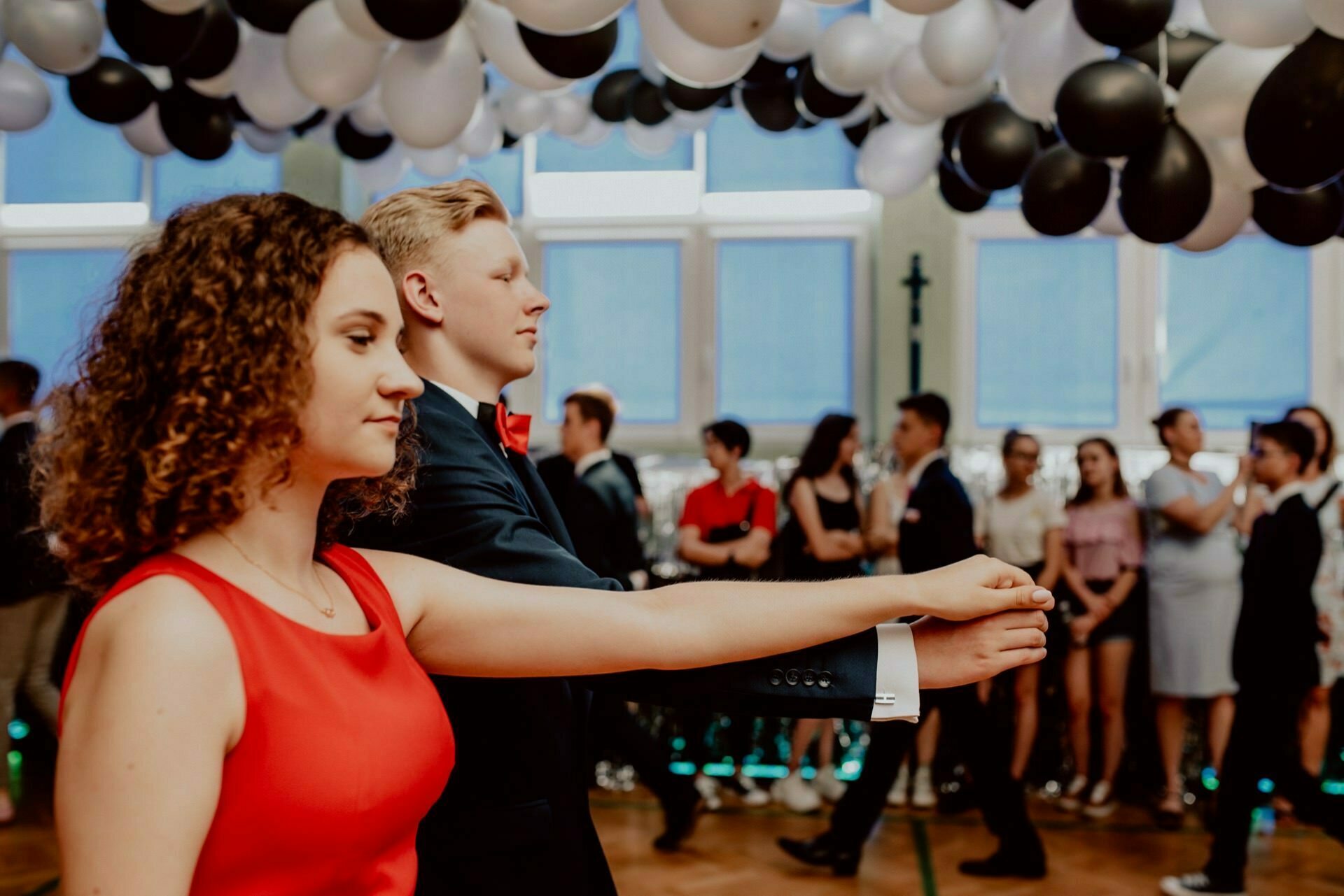 A young woman in a red dress and a young man in a black suit with a red bow tie dance together in a decorated room with black and white balloons on the ceiling. A group of people in the background watch the dance, perfectly capturing the essence of event photography. 