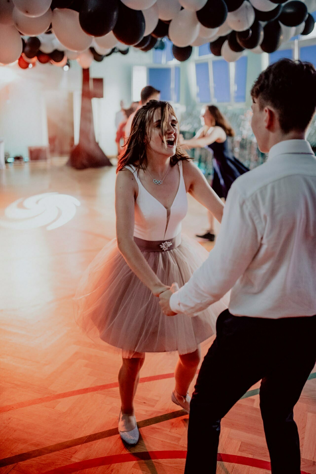 A young woman in a white top and pink tulle skirt vigorously dances with a young man in a white shirt at a party, under a ceiling decorated with black and white balloons. There are other people dancing in the background of this well-lit room, perfectly captured by our event photographer Warsaw. 