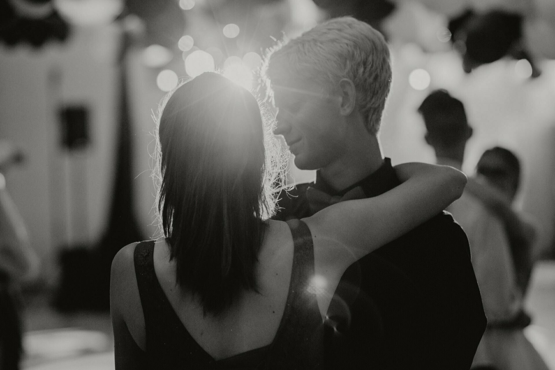 Black and white photo of a couple dancing close, with the woman's arm slung over the man's shoulder. They stand facing each other, lit by soft light in the background, creating a romantic atmosphere. This event photography beautifully captures their intimate bond.  