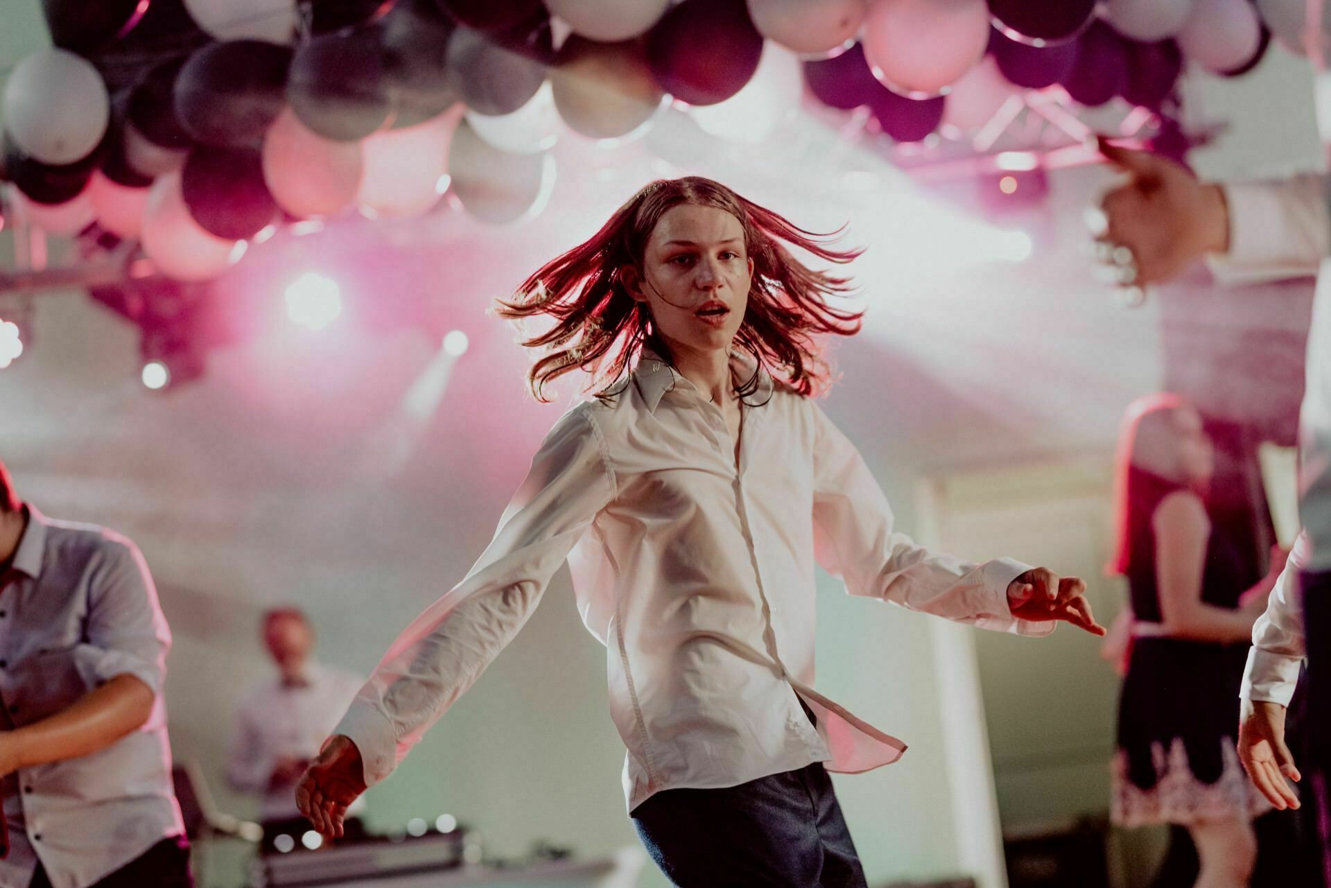 A person with long hair, wearing a white shirt, dances vigorously under a ceiling filled with black and white balloons. The background is lit with pinkish lights, and the other dancers are partially visible - which really captures the essence of *event photography* in this vibrant scene. 