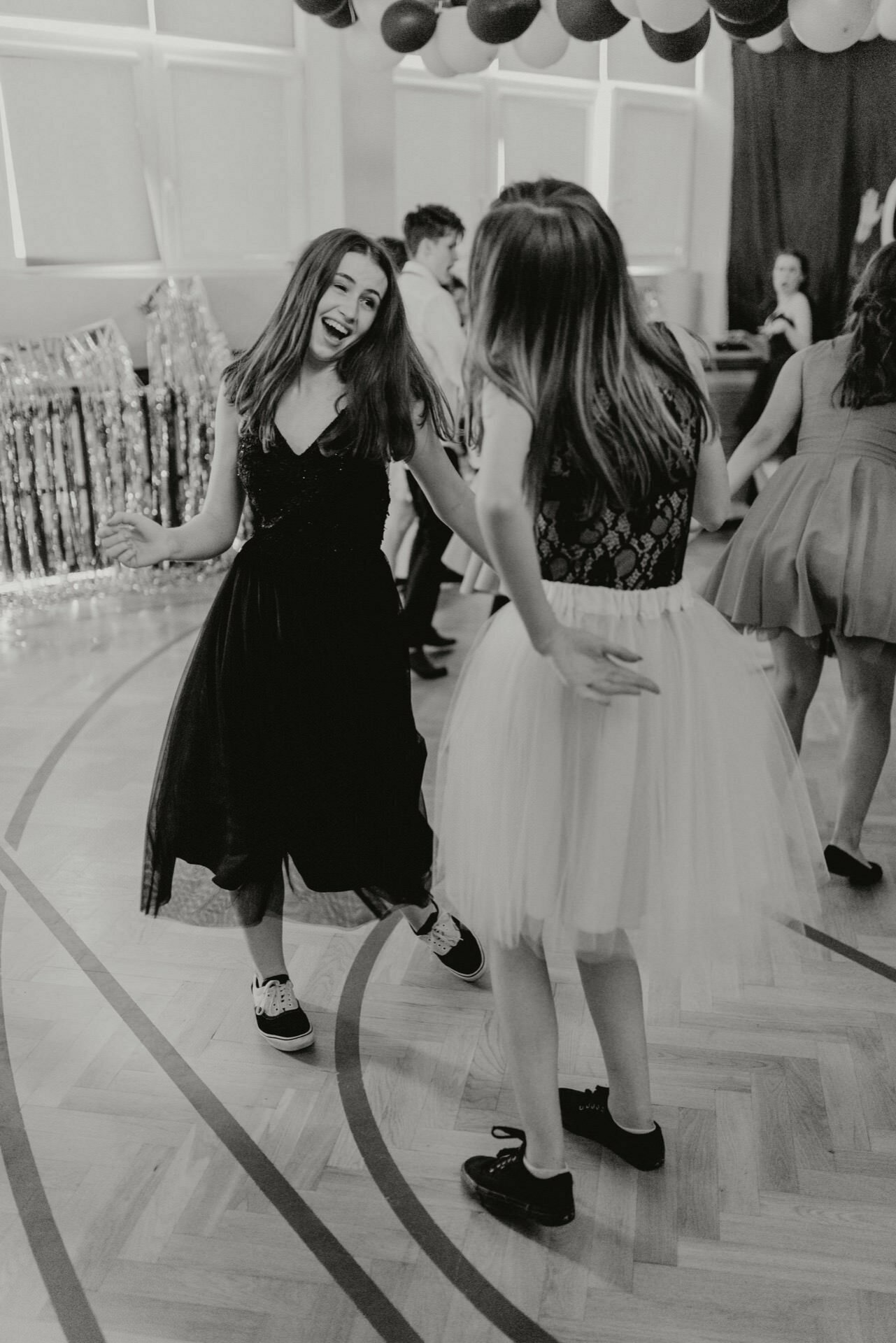 Black and white image of two girls dancing happily at a party. One is wearing a dark dress and the other a light-colored dress with a tulle skirt. They are surrounded by other people and festive decorations, including hanging balloons and shimmering streamers - perfect for any *event photography*.  